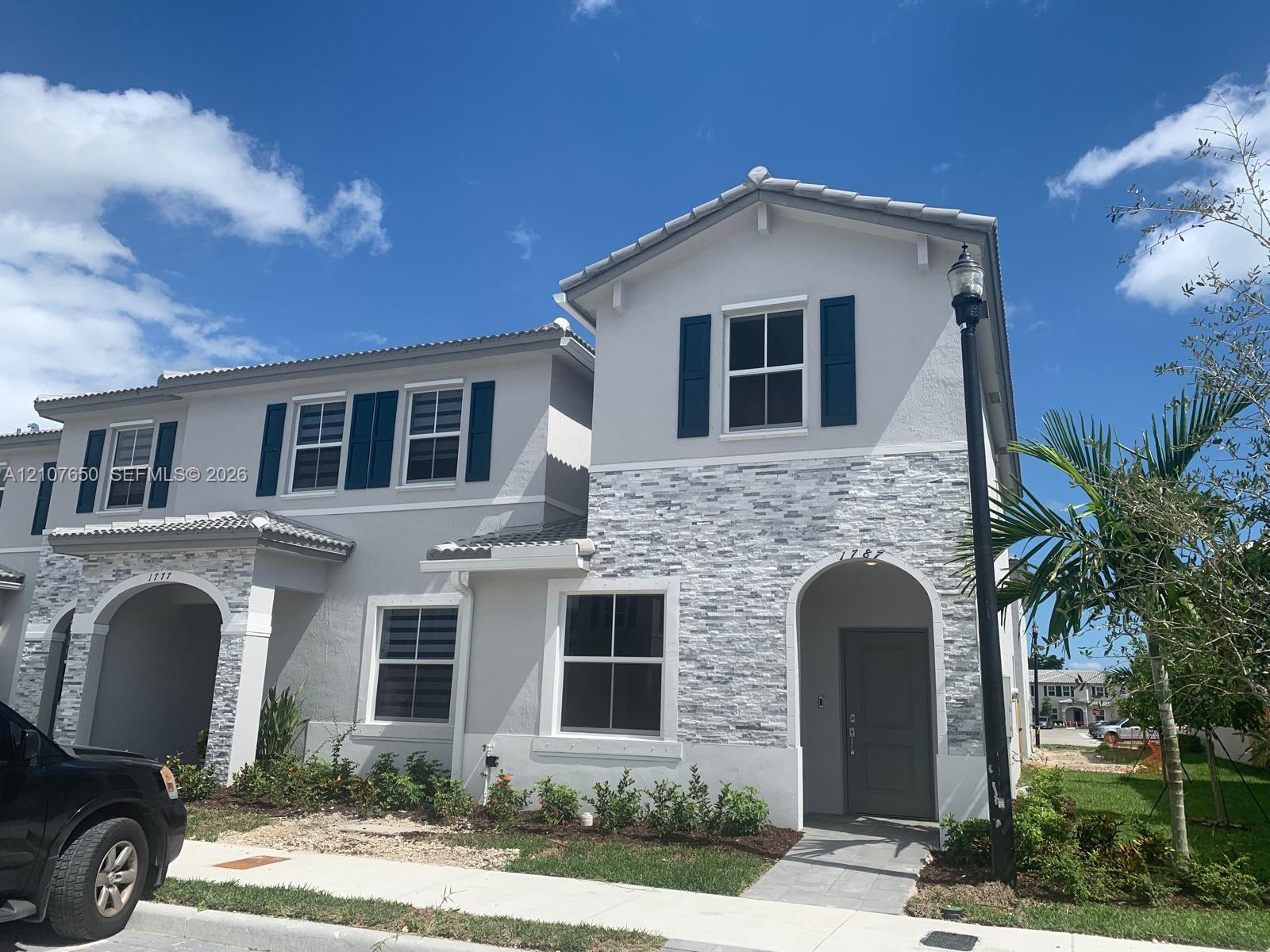 Two-story exterior featuring a light gray stucco facade with stone veneer accents, arched entryways, dark-toned shutters, and a tiled roof