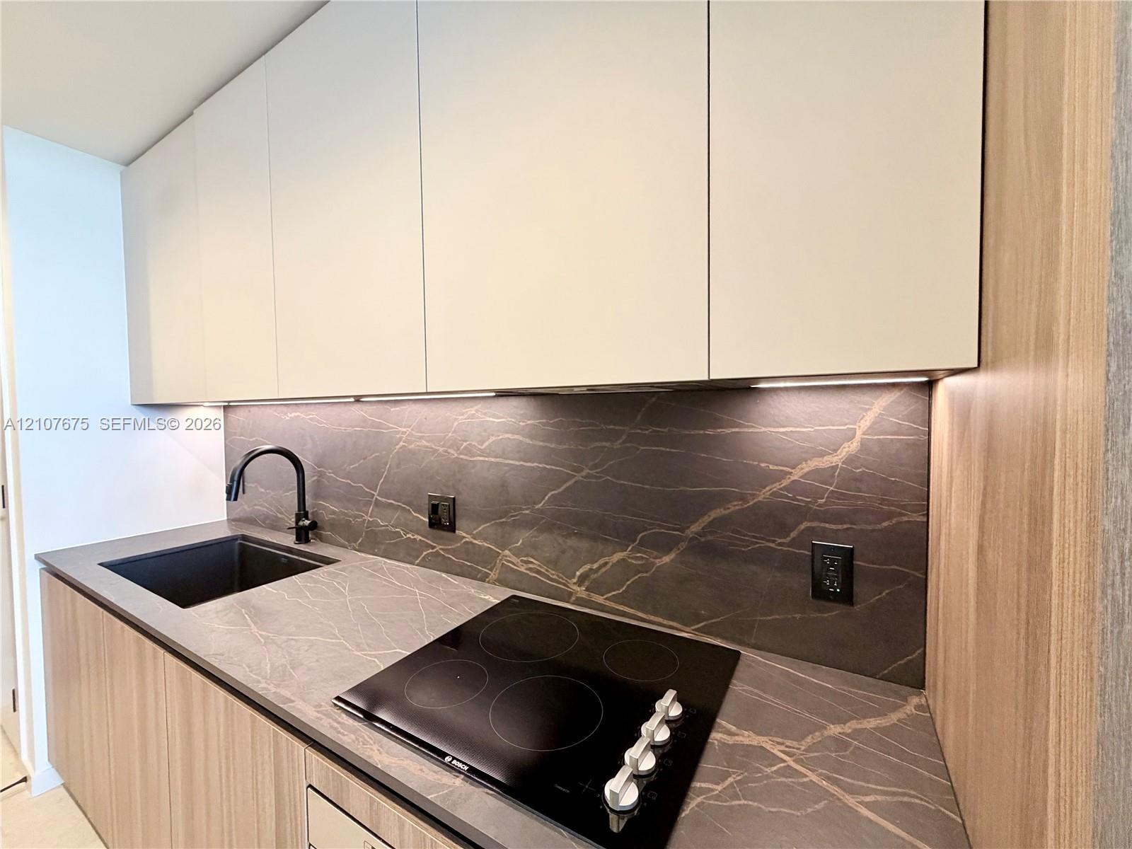 Sleek kitchen featuring flat-panel cabinetry, stone-finish countertops and backsplash, integrated four-burner induction cooktop, and a matte black gooseneck faucet