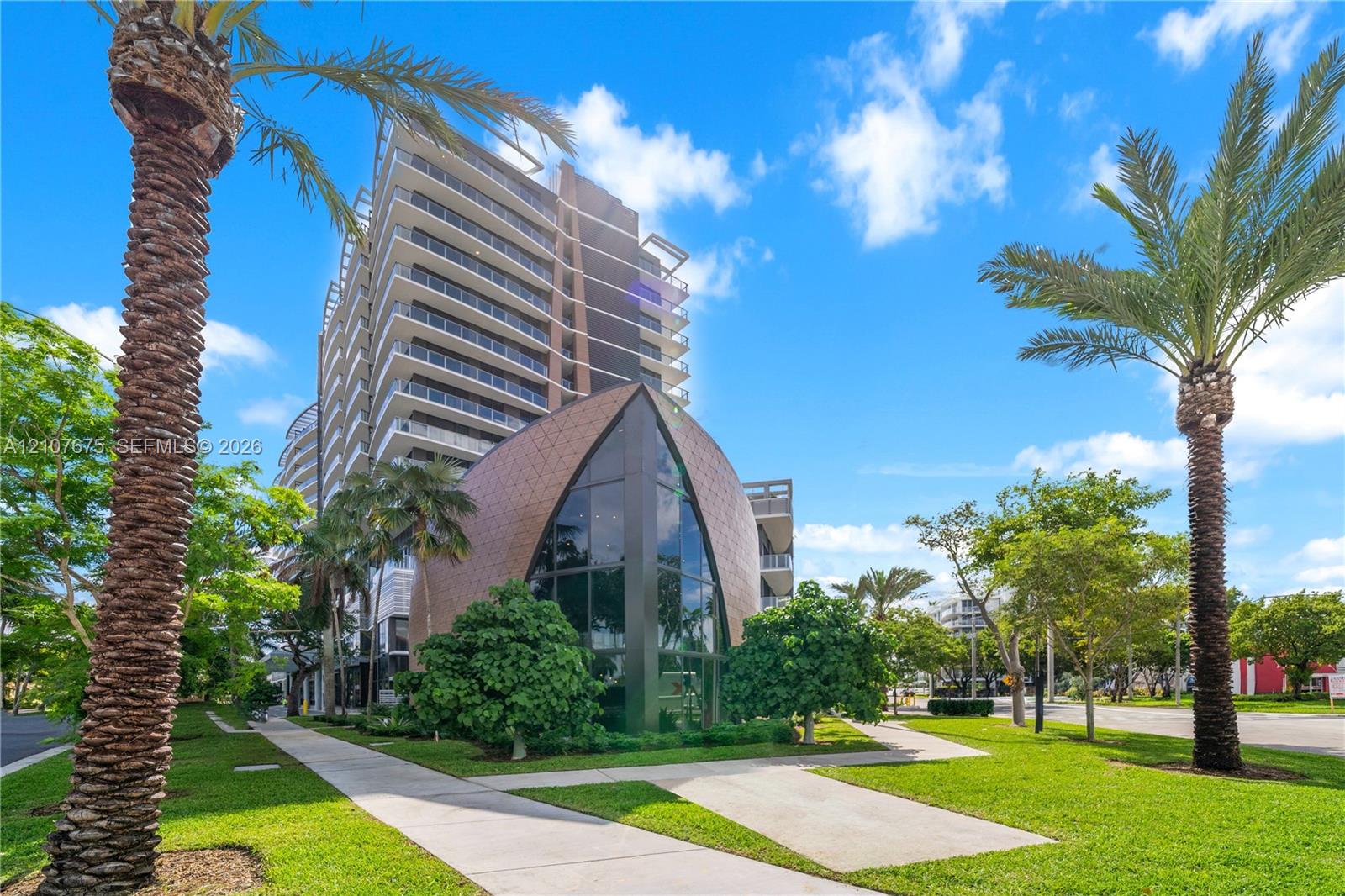 High-rise residential exterior featuring expansive glass curtain walls, private balconies, a unique arched architectural structure, and manicured grounds with palm trees High-rise residential exterior featuring expansive glass curtain walls, private balconies, a unique arched architectural structure, and manicured grounds with palm trees