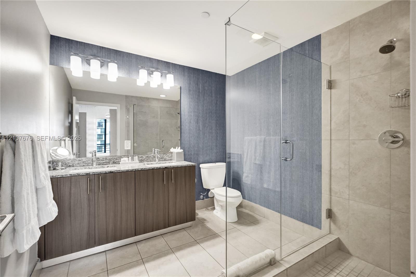 Bathroom featuring a dual-sink vanity with stone countertop, frameless glass-enclosed shower, large-format tile flooring, and integrated wall lighting