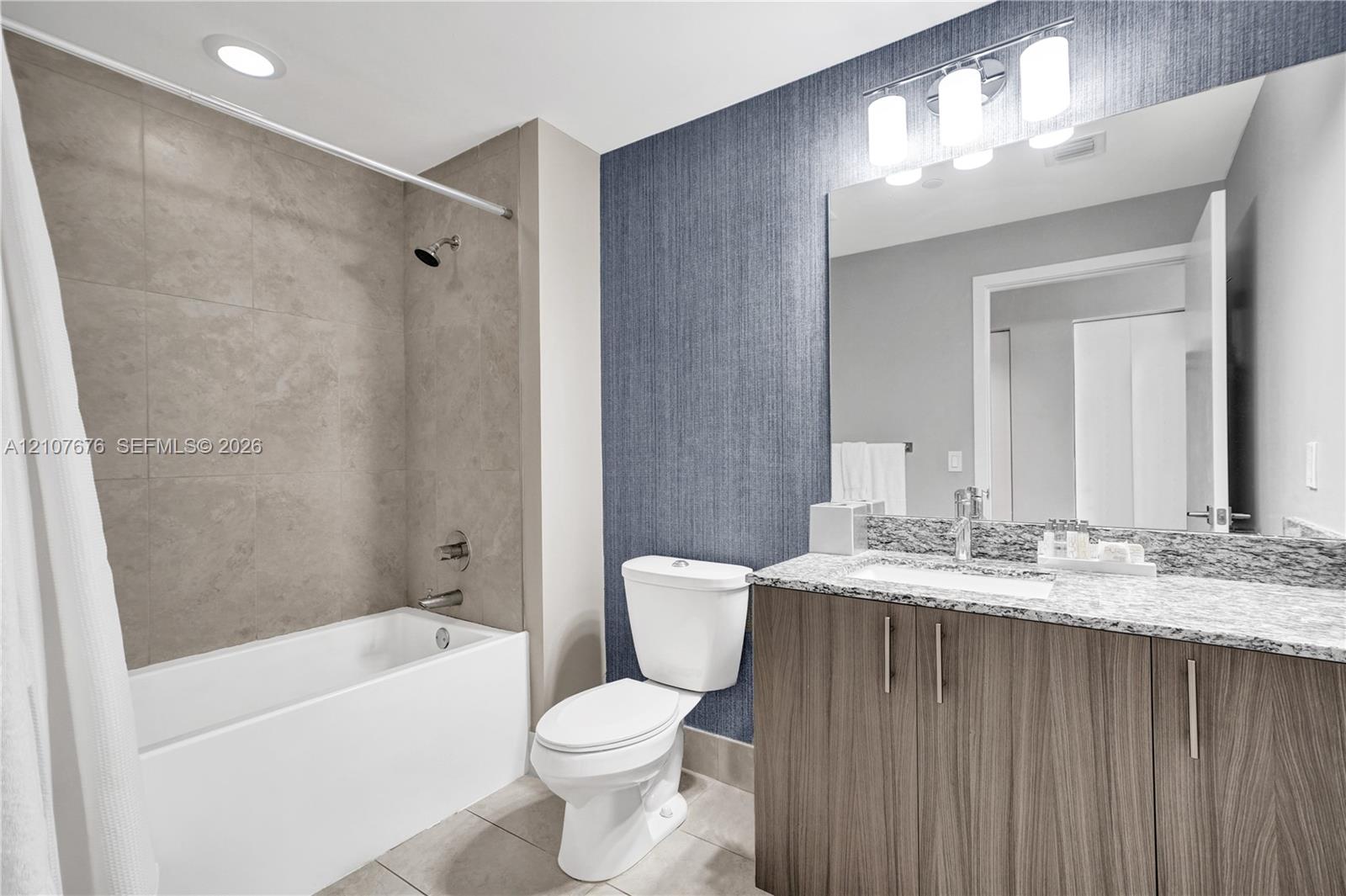 Bathroom featuring a vanity with wood-finish cabinetry, a stone-composite countertop with an integrated sink, textured navy blue wall covering, a large wall-mounted mirror, and a tub-shower combination with floor-to-ceiling tile surround