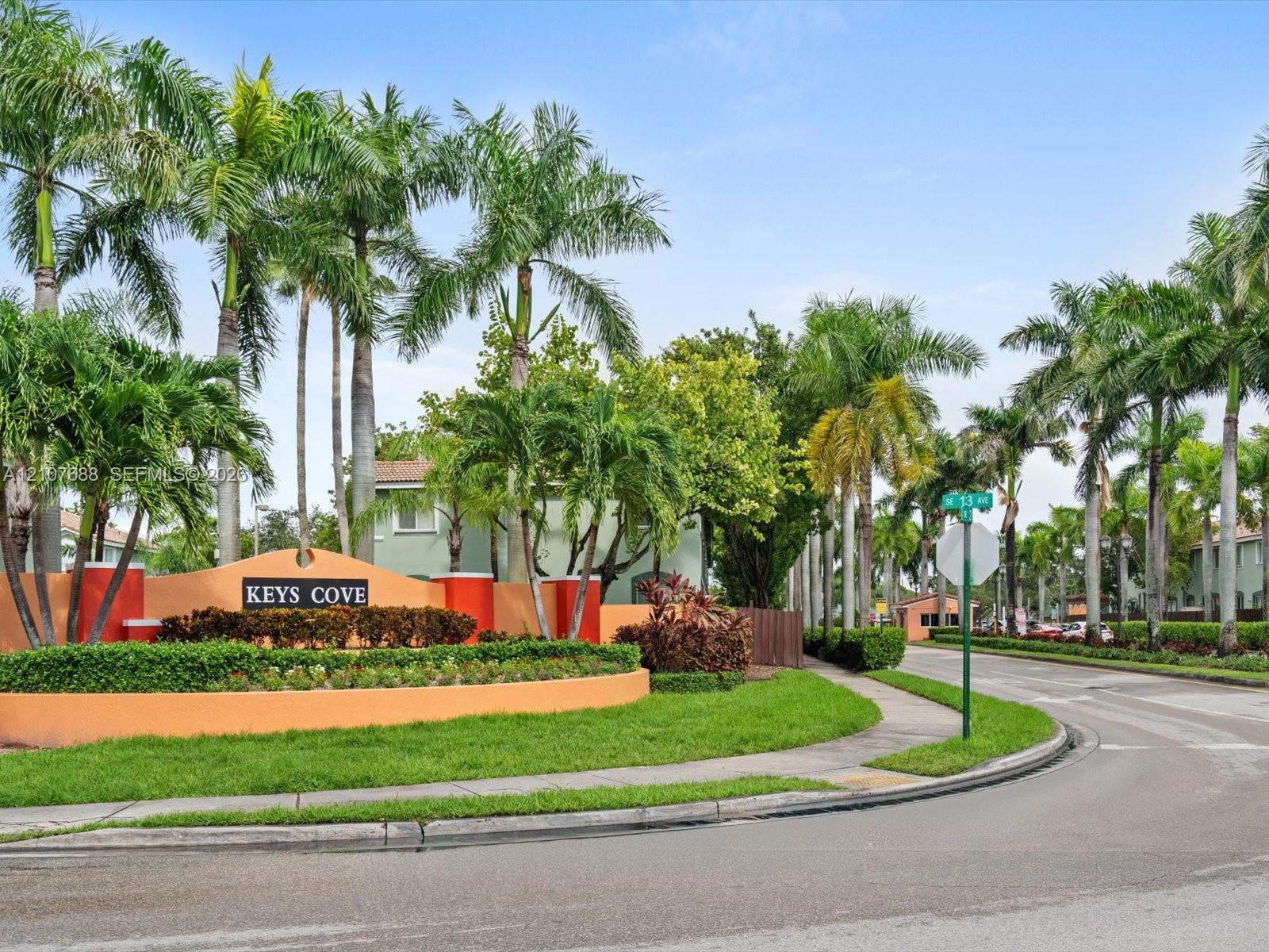 Community entrance monument with stucco finish, lush landscaping, mature palm trees, manicured lawn, and asphalt paved thoroughfare