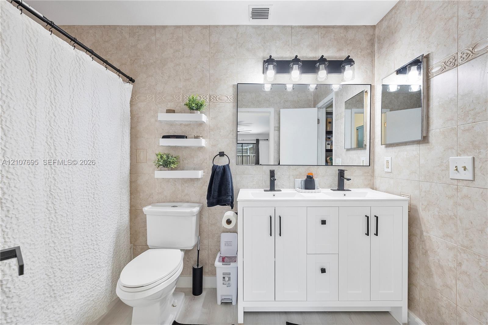 Double-sink vanity with white cabinetry, matte black hardware, and rectangular undermount sinks, complemented by a wide vanity mirror, four-light wall fixture, and neutral-toned wall tiling