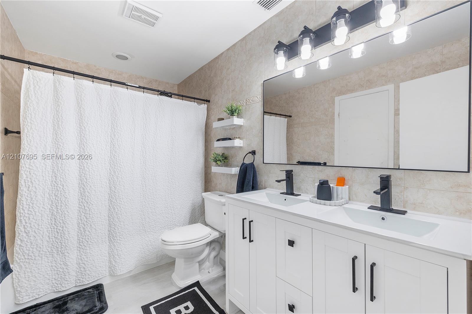 Dual-sink vanity with white cabinetry, matte black hardware, stone-surface countertop, and rectangular undermount sinks, complemented by a large-format mirror with integrated multi-bulb vanity lighting and neutral tile wall cladding