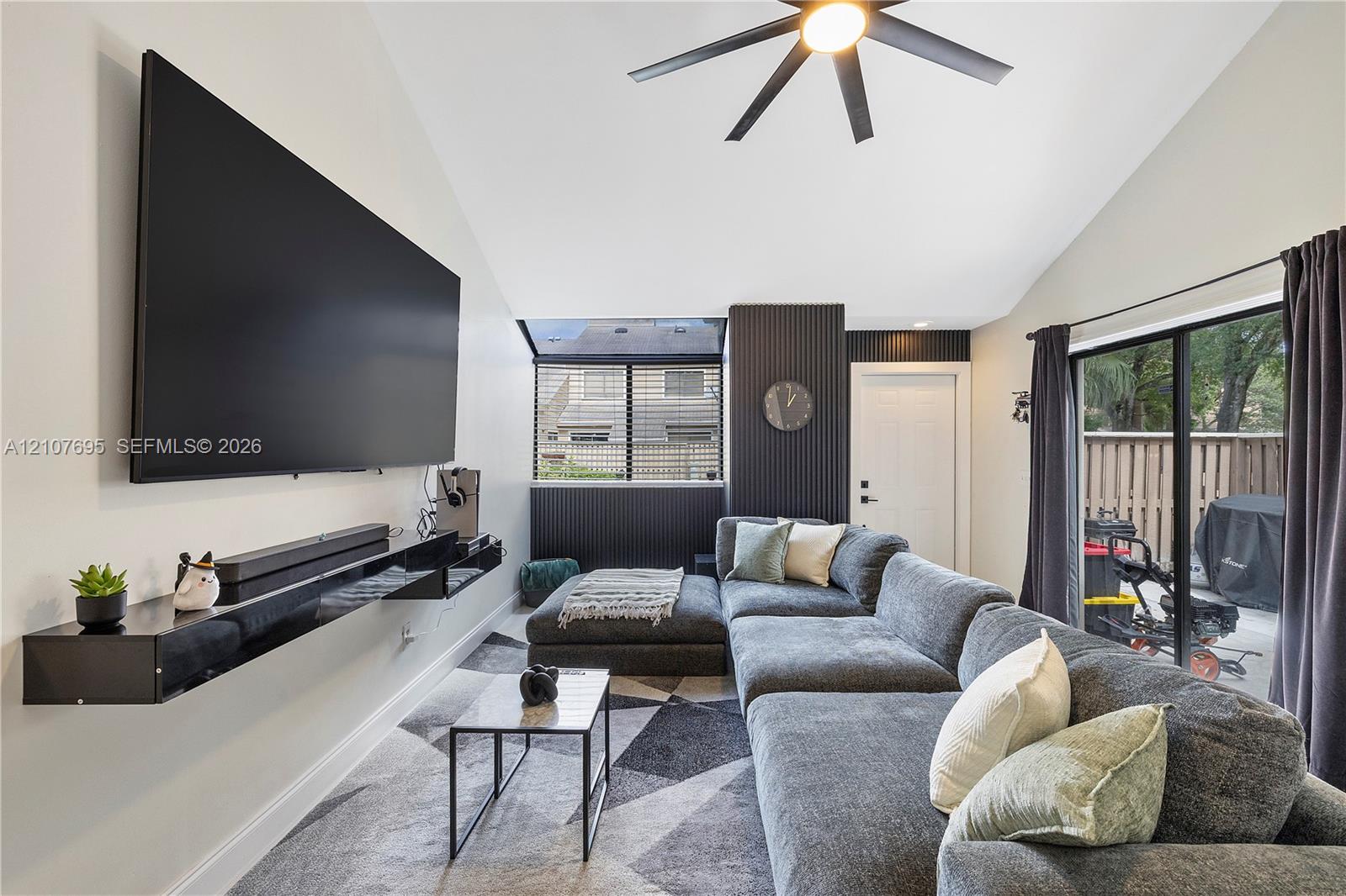 Vaulted ceilings, dark accent wall with vertical slat detailing, matte black ceiling fan with integrated lighting, floating black media console, and sliding glass doors