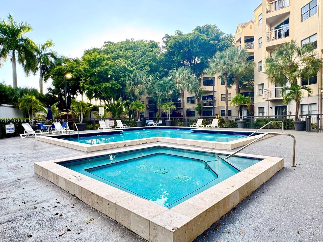 In-ground swimming pool and spa featuring stone tile coping, surrounded by a paved deck and mature palm landscaping