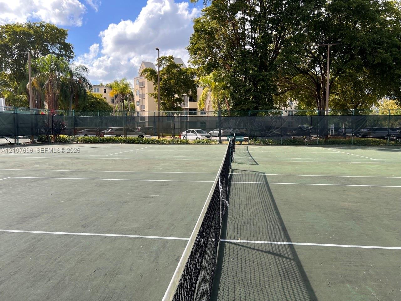 Hard-court tennis facility featuring a central net, perimeter fencing, and mature trees