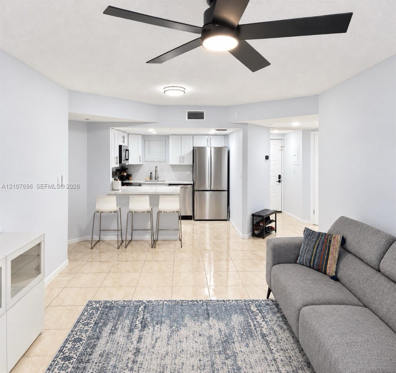Open-concept interior featuring light tile flooring, matte black ceiling fan with integrated lighting, stainless steel refrigerator, white cabinetry, and a peninsula counter