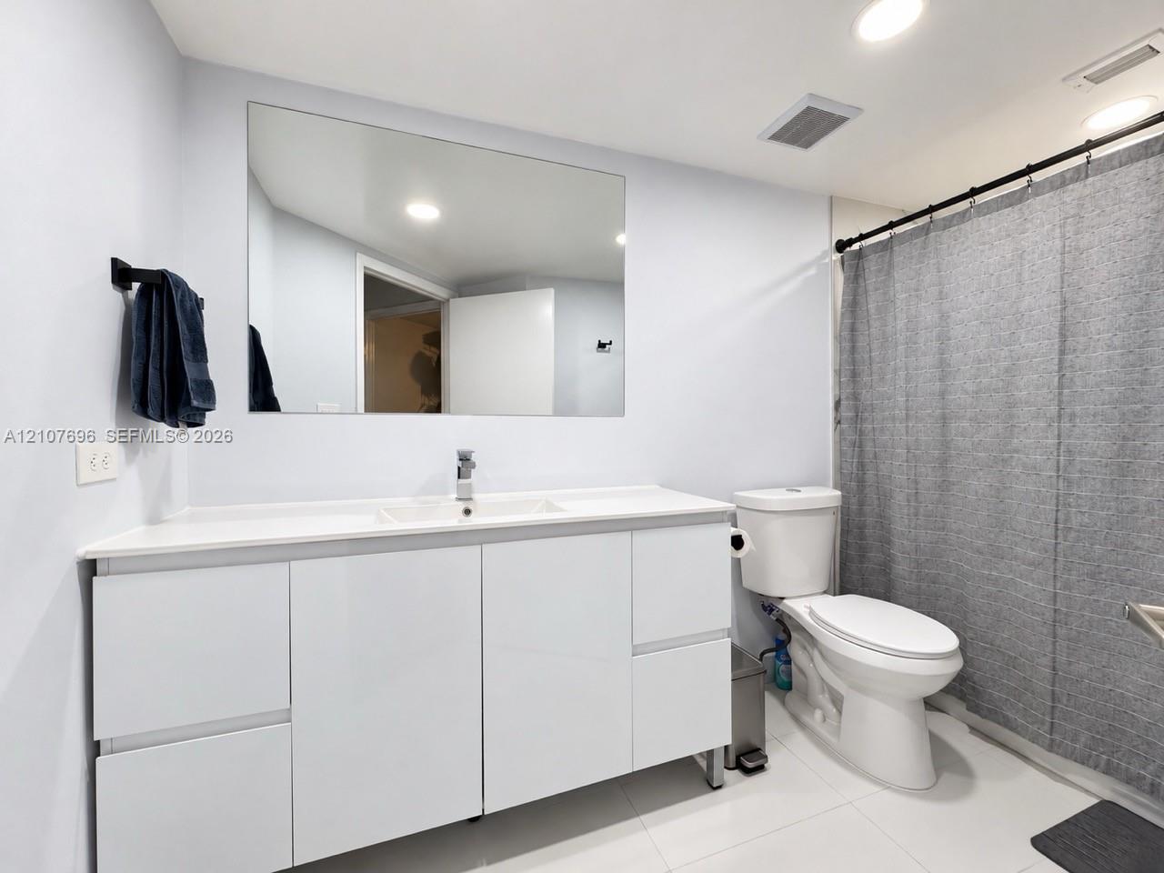 Modern bathroom vanity with high-gloss white cabinetry, integrated sink, and polished chrome faucet featuring a large frameless wall mirror, recessed lighting, and tile flooring