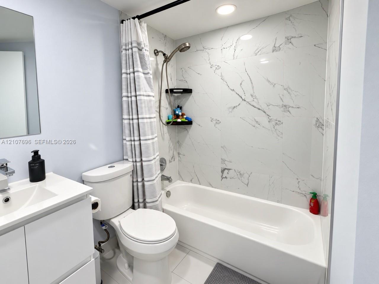 Bathroom featuring white vanity with integrated sink, porcelain tile surround with veined finish, chrome handheld shower fixture, and recessed lighting