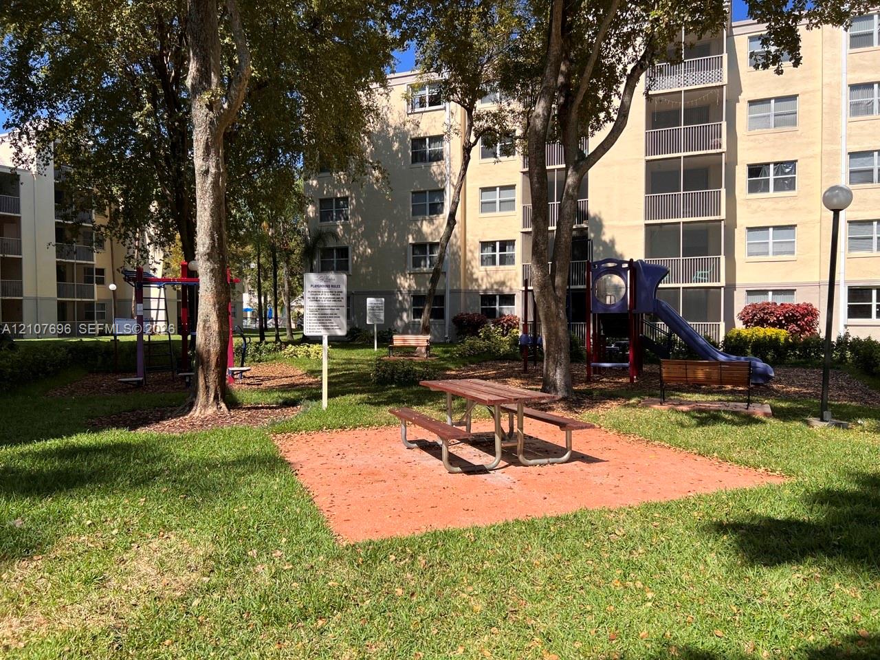 Common area featuring a recreational playground, manicured lawn, mature trees, and a fixed metal picnic table