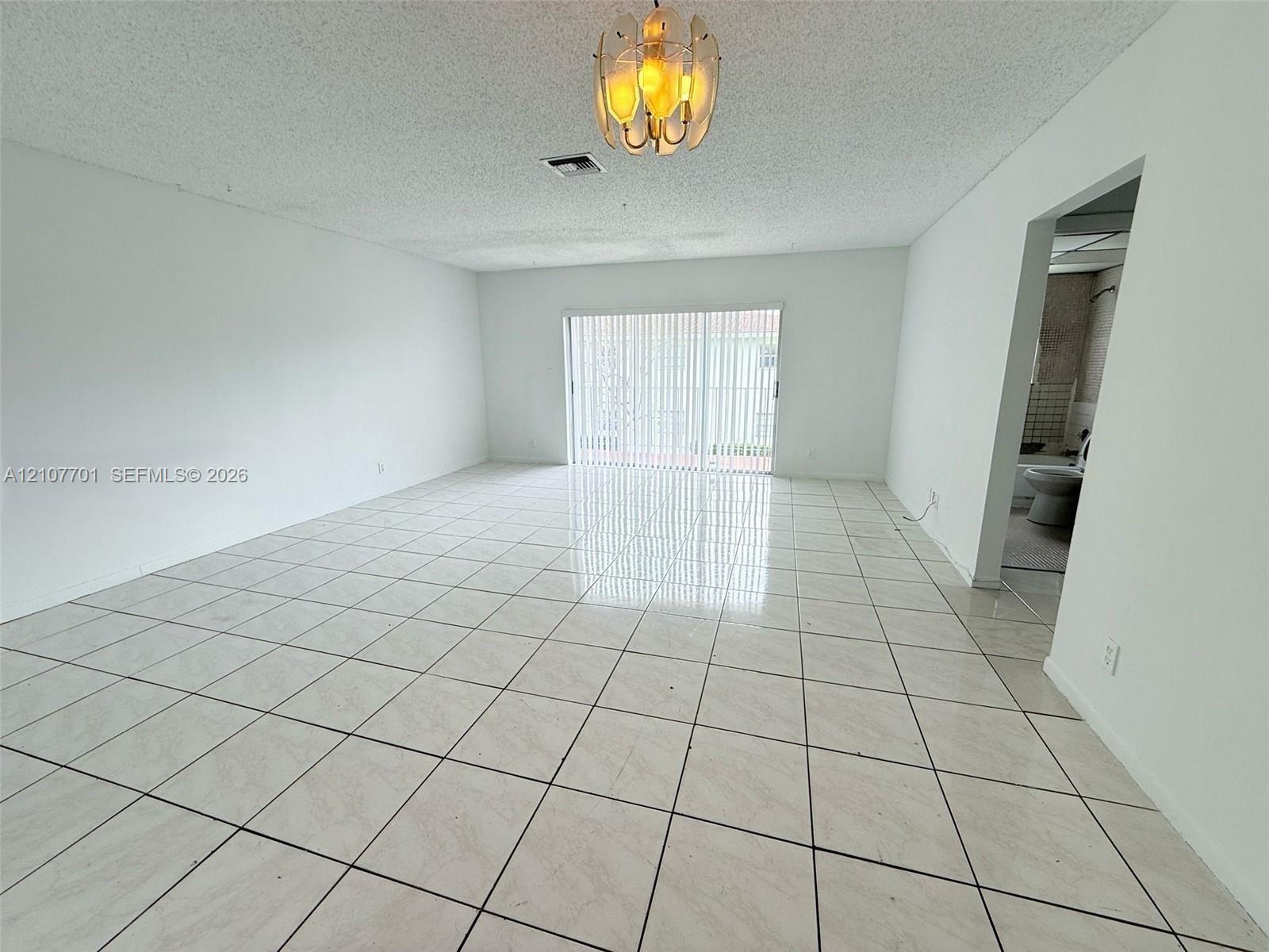 Open-concept area featuring light-toned tile flooring, textured ceiling, glass sliding doors with vertical blinds, and a multi-bulb flush-mount light fixture