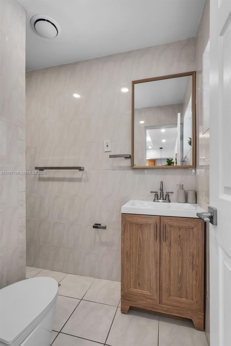 Bathroom featuring a wood-finish vanity with a rectangular white sink, metallic faucet, framed wall mirror, and floor-to-ceiling tile work