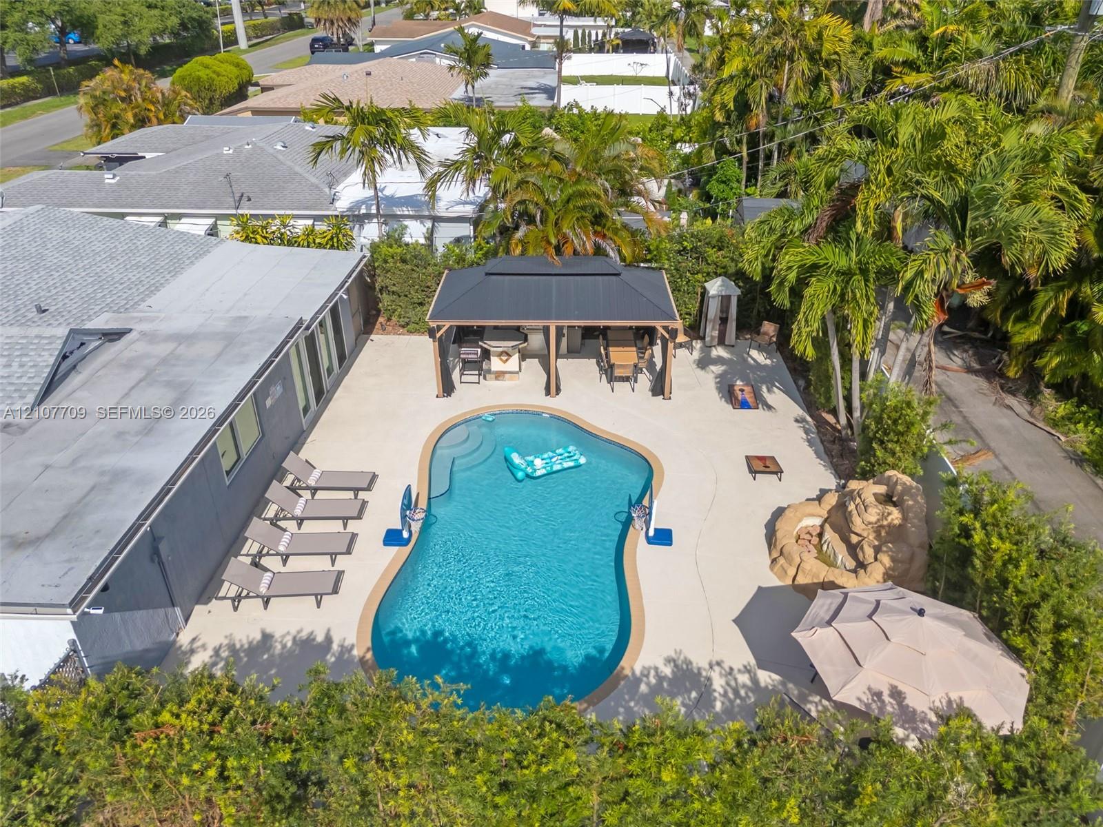 Backyard area featuring an inground pool with a curvilinear edge, a covered gazebo, a decorative stone-finish rock feature, and expansive concrete hardscaping surrounded by mature perimeter landscaping