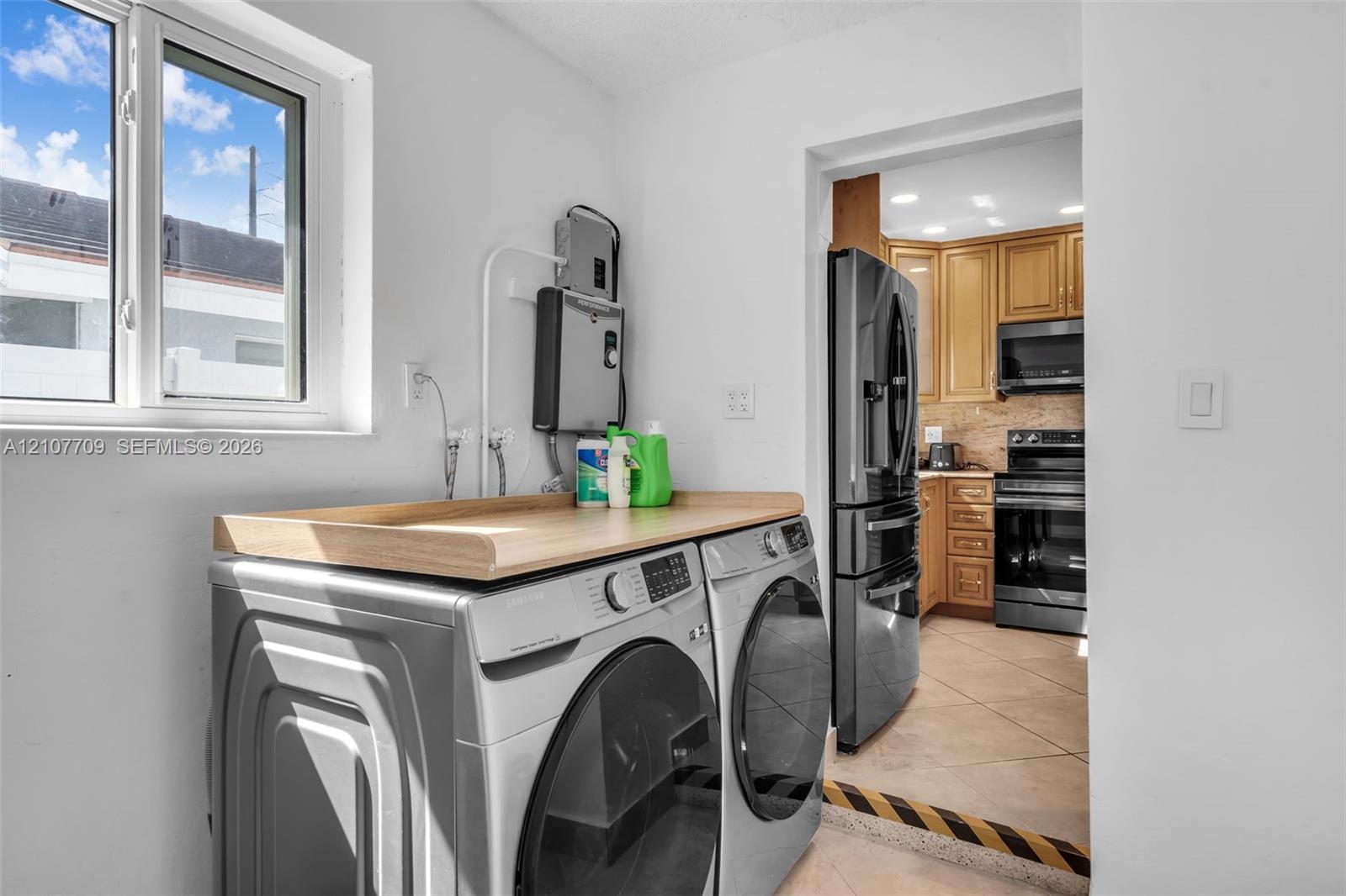 Laundry space featuring a wooden folding counter, vinyl-frame window, and tiled flooring extending into the kitchen area with wood-finish cabinetry and stainless steel appliances