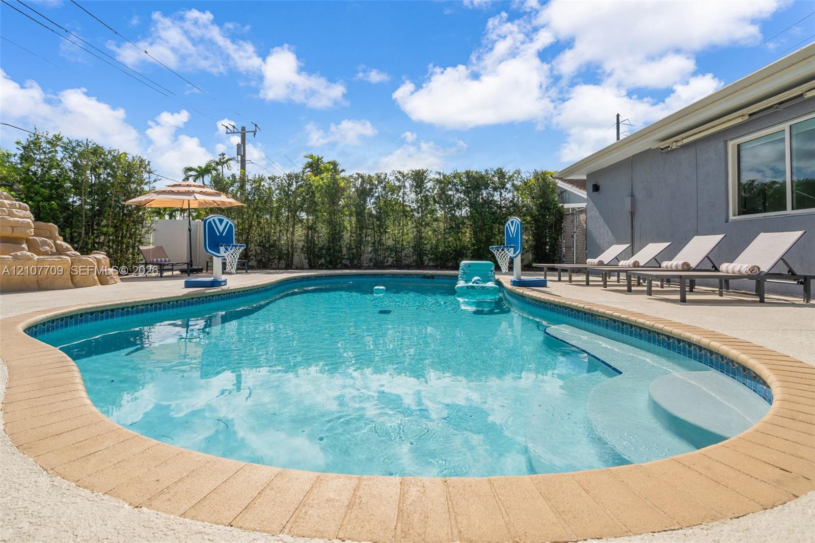 In-ground swimming pool featuring a tan brick border, built-in steps, and a decorative stone water feature