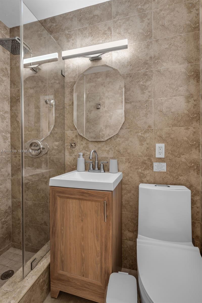 Bathroom featuring full-height neutral wall tile, wood-finish vanity with a rectangular sink, glass-enclosed shower with rainfall showerhead, and a wall-mounted light fixture