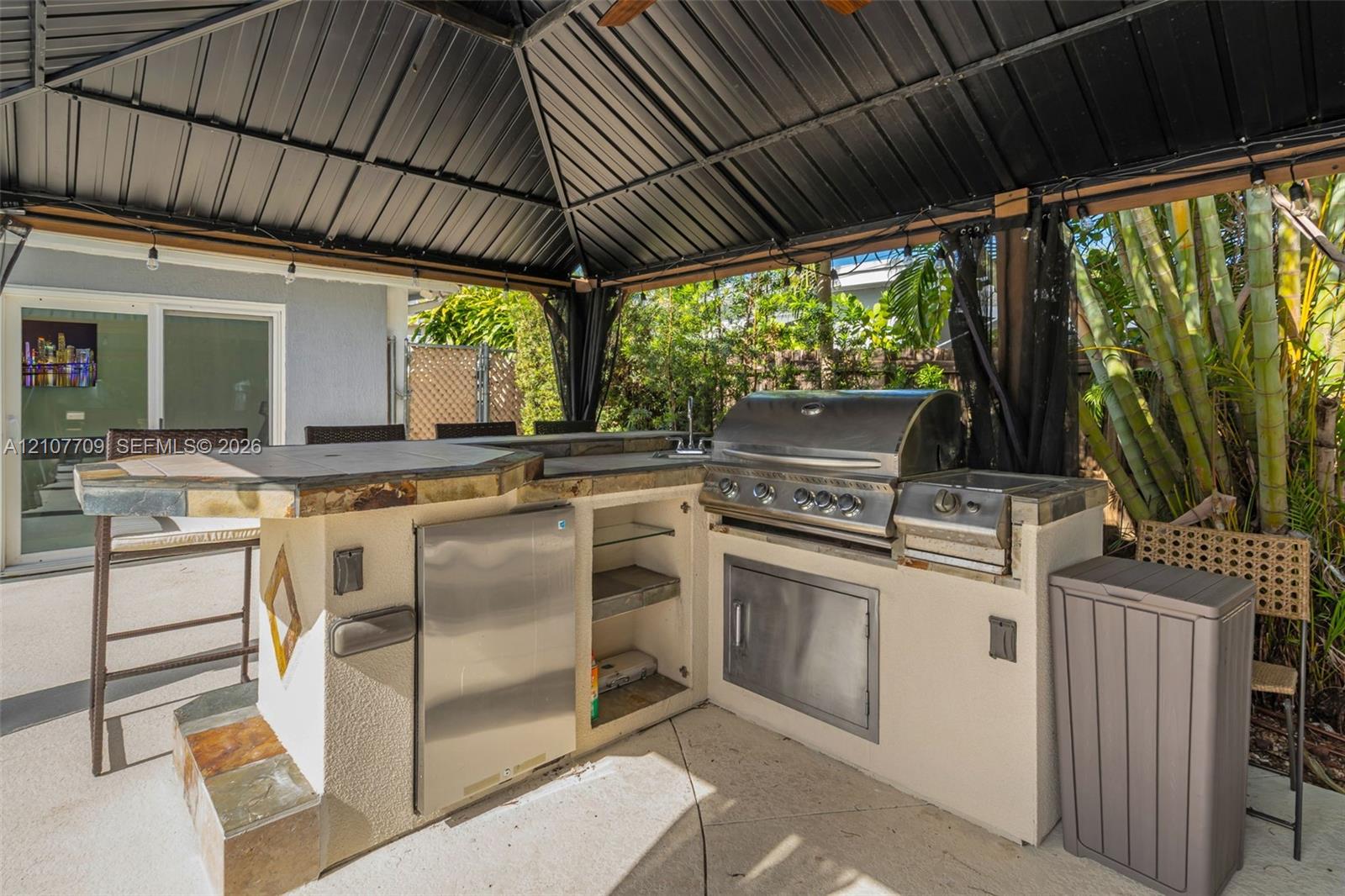 Outdoor kitchen featuring a stone-clad island with a counter, stainless steel built-in grill, stainless steel refrigerator, and protective metal-roofed gazebo