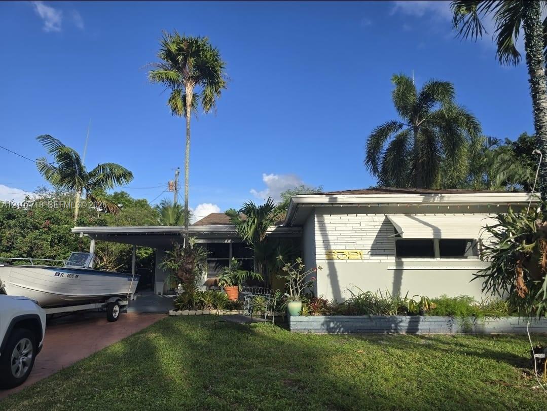 Single-story residence featuring a painted masonry facade, metal awning window coverings, a covered carport, and established tropical landscaping Single-story residence featuring a painted masonry facade, metal awning window coverings, a covered carport, and established tropical landscaping
