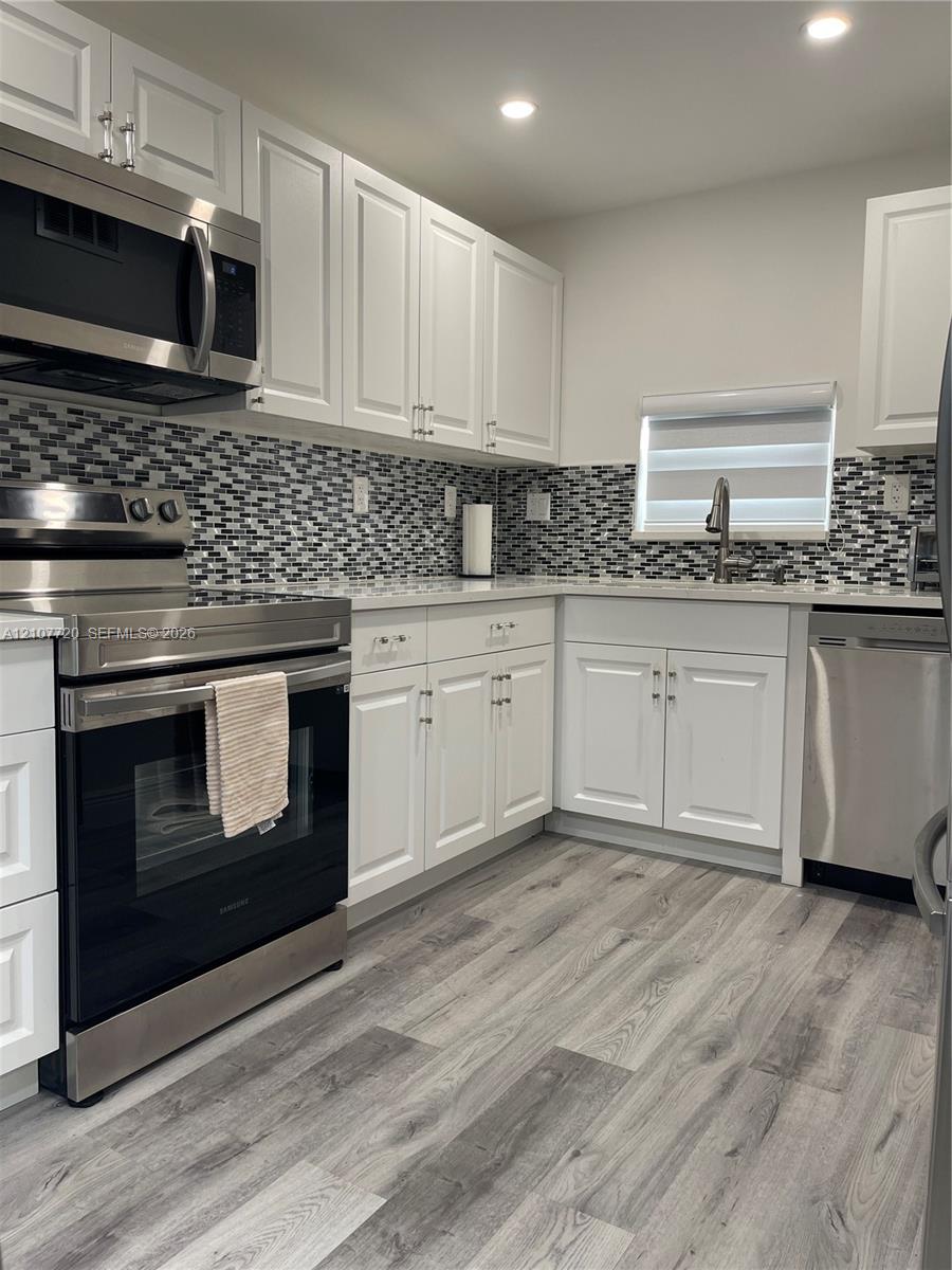 Kitchen featuring white shaker-style cabinetry, stainless steel appliances, multi-tonal mosaic tile backsplash, stone-finish countertops, and wood-finish flooring Kitchen featuring white shaker-style cabinetry, stainless steel appliances, multi-tonal mosaic tile backsplash, stone-finish countertops, and wood-finish flooring