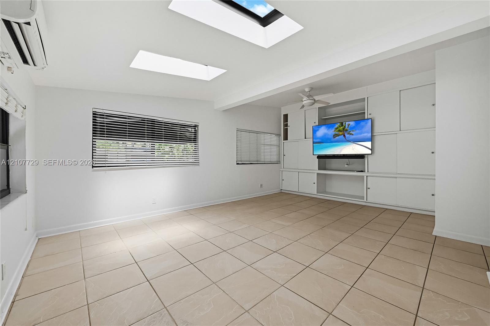 Large room featuring neutral tile flooring, dual skylights, custom built-in cabinetry, horizontal window blinds, and a ceiling fan