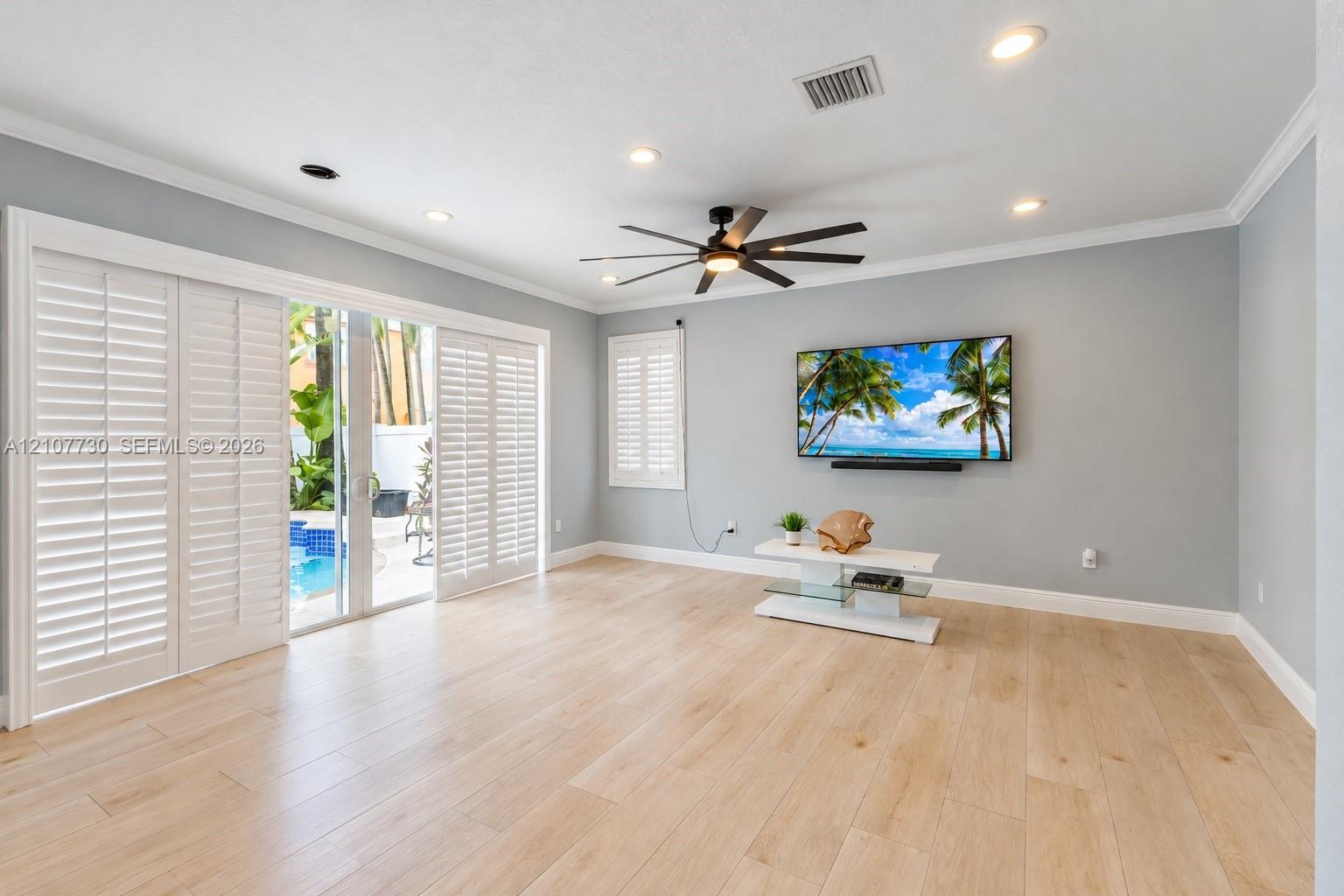 Open-concept room featuring light wood-finish flooring, recessed lighting, a modern ceiling fan, sliding glass doors with plantation shutters, and crown molding