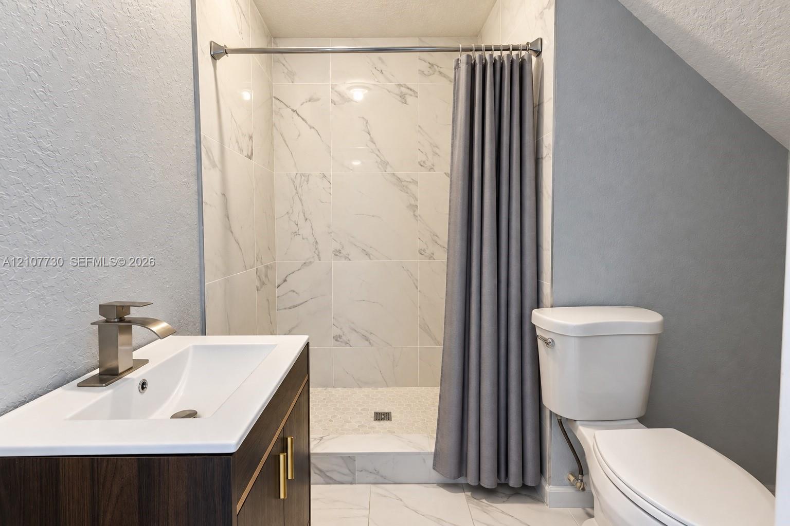 Bathroom featuring a wood-finish vanity with a rectangular sink, matte metallic faucet, large-format tiled shower surround with stone-pattern detail, and stone-finish floor tiling