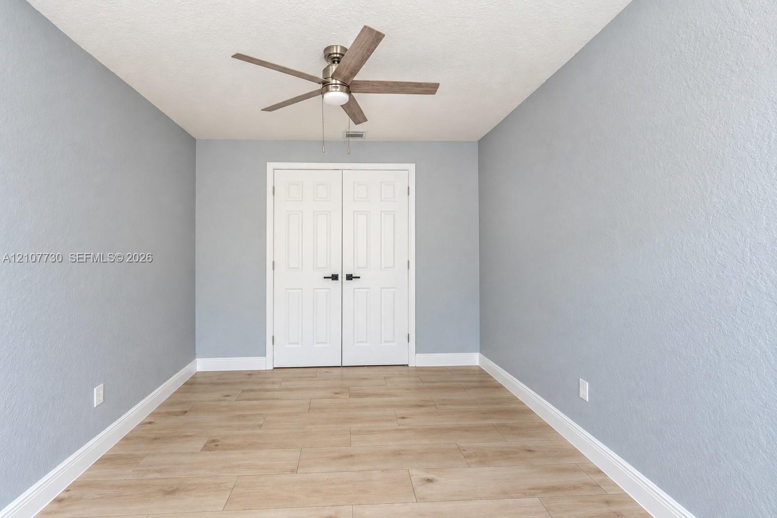 Room featuring wood-finish tile flooring, light blue textured walls, dual-panel closet doors with matte black hardware, and a ceiling fan with integrated lighting