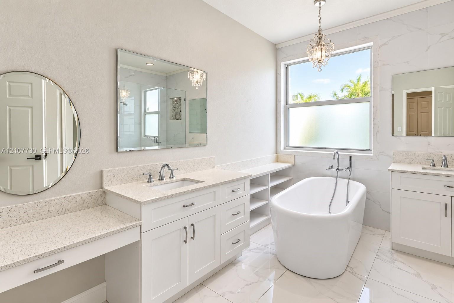 Freestanding soaking tub with chrome wall-mounted faucet, dual white vanities with stone countertops, integrated open shelving, and large-format tile flooring
