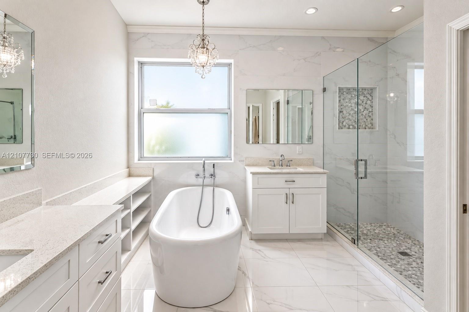 Bathroom featuring a freestanding soaking tub, walk-in glass-enclosed shower with a stone-tile floor, dual white vanities with solid-surface countertops, and decorative pendant lighting