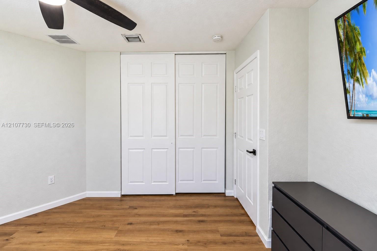 Neutral-toned interior featuring wood-finish flooring, sliding dual-panel closet doors, and a ceiling fan with integrated lighting