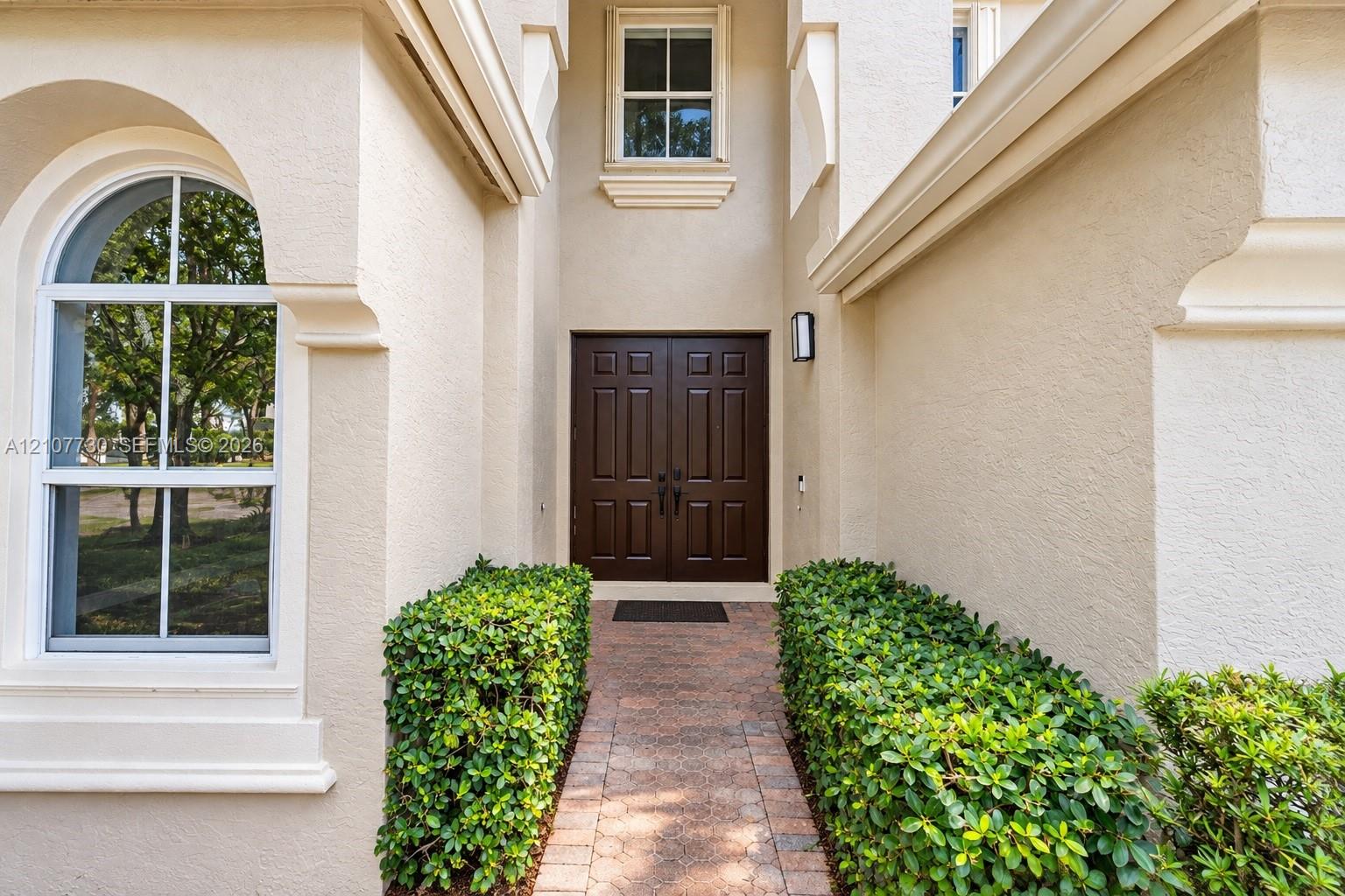 Stucco exterior entrance featuring a double-door entry, arched window, stone-paver walkway, and manicured shrubbery