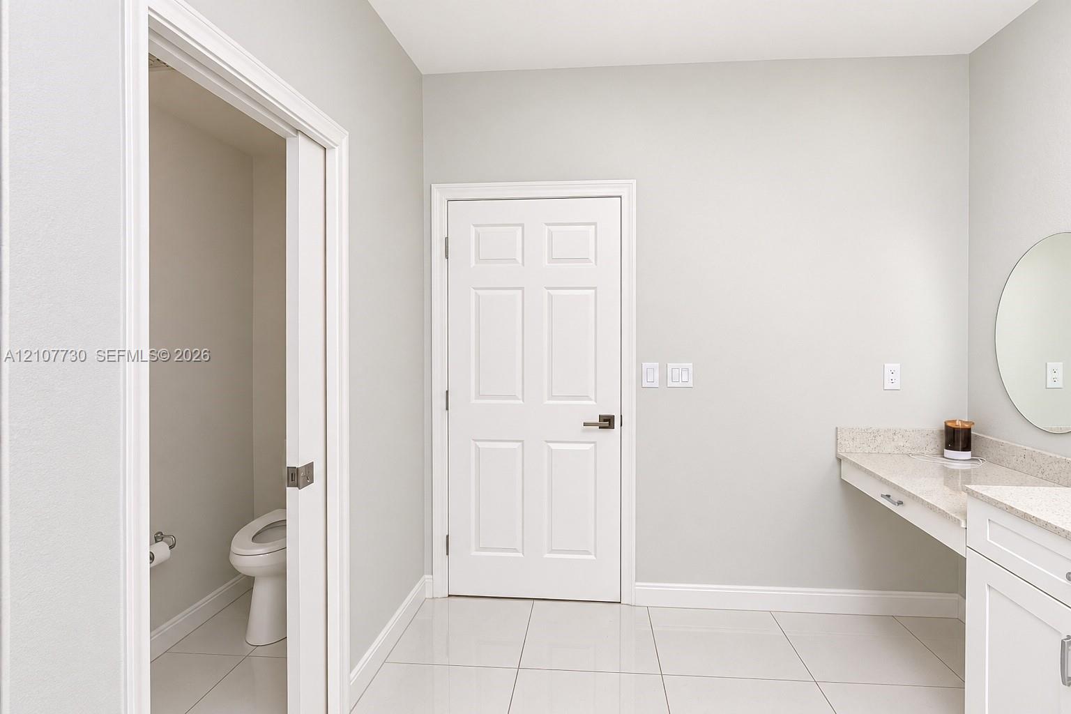 Light-toned tile flooring, white six-panel door, built-in vanity with stone-composite countertop, and wall-mounted circular mirror