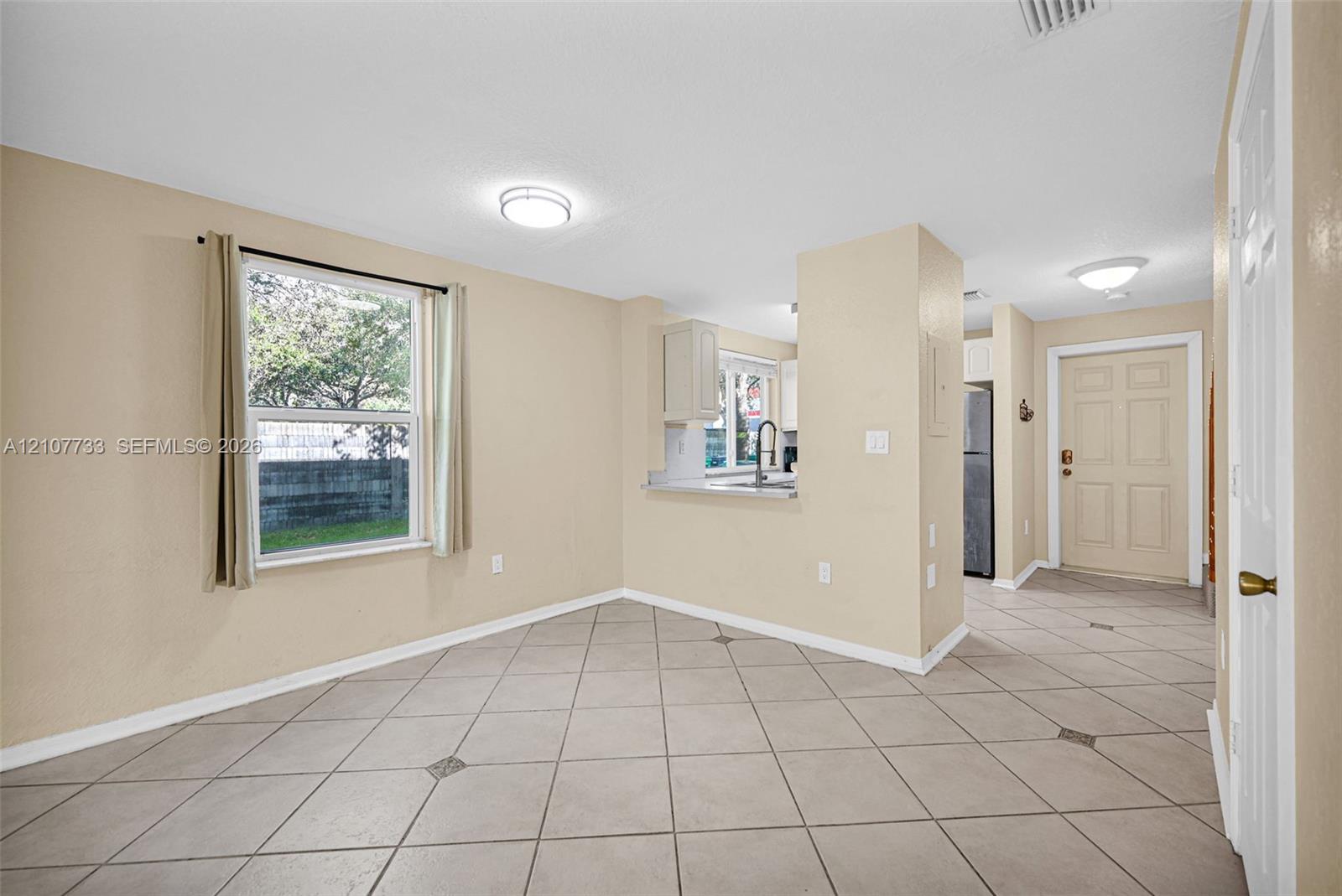 Tile flooring set on a diagonal pattern, neutral wall color, oversized window, and kitchen pass-through with a light-toned countertop