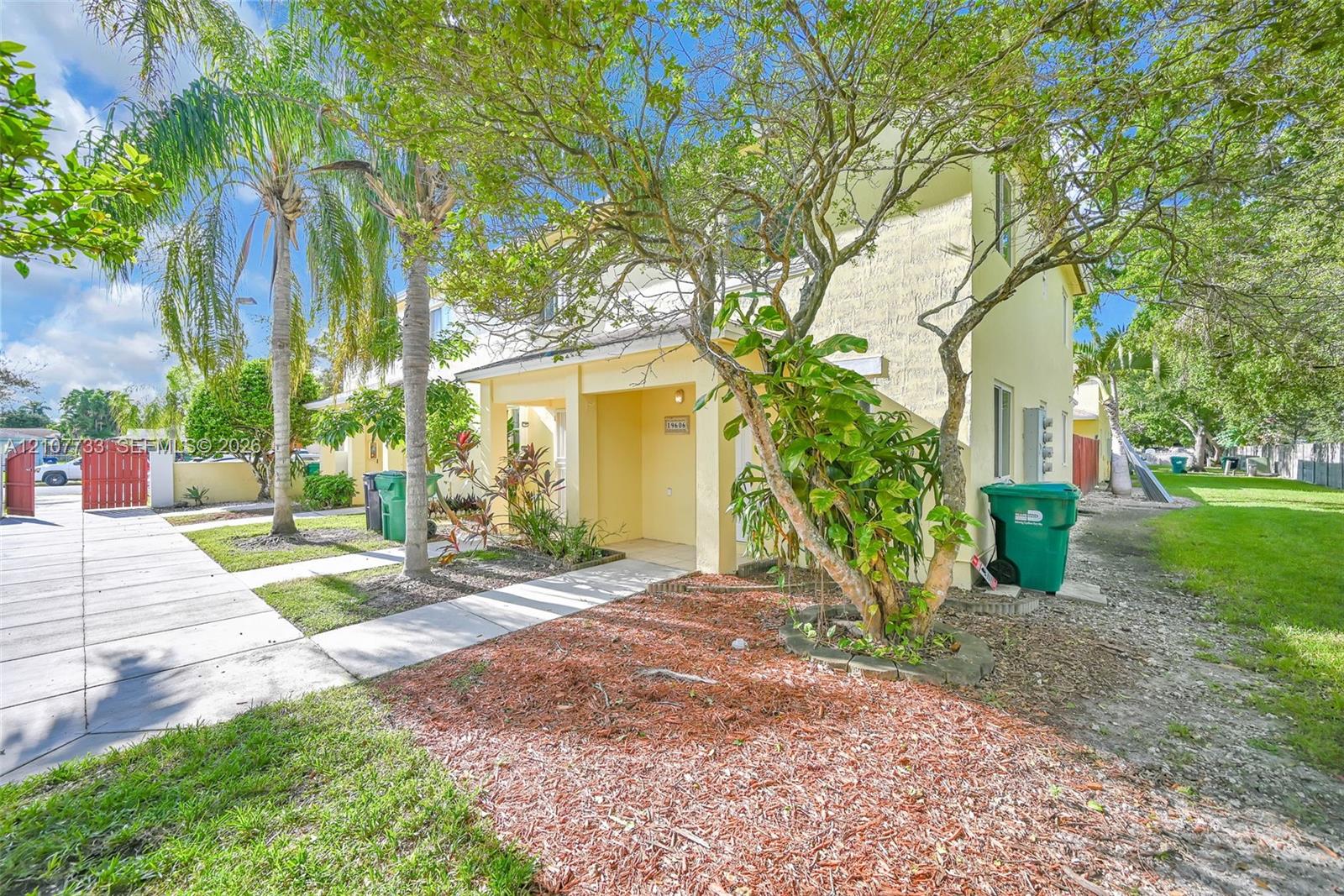 Two-story exterior featuring a stucco facade, covered entryway, concrete walkway, and landscaped garden area with mulch beds and palm trees