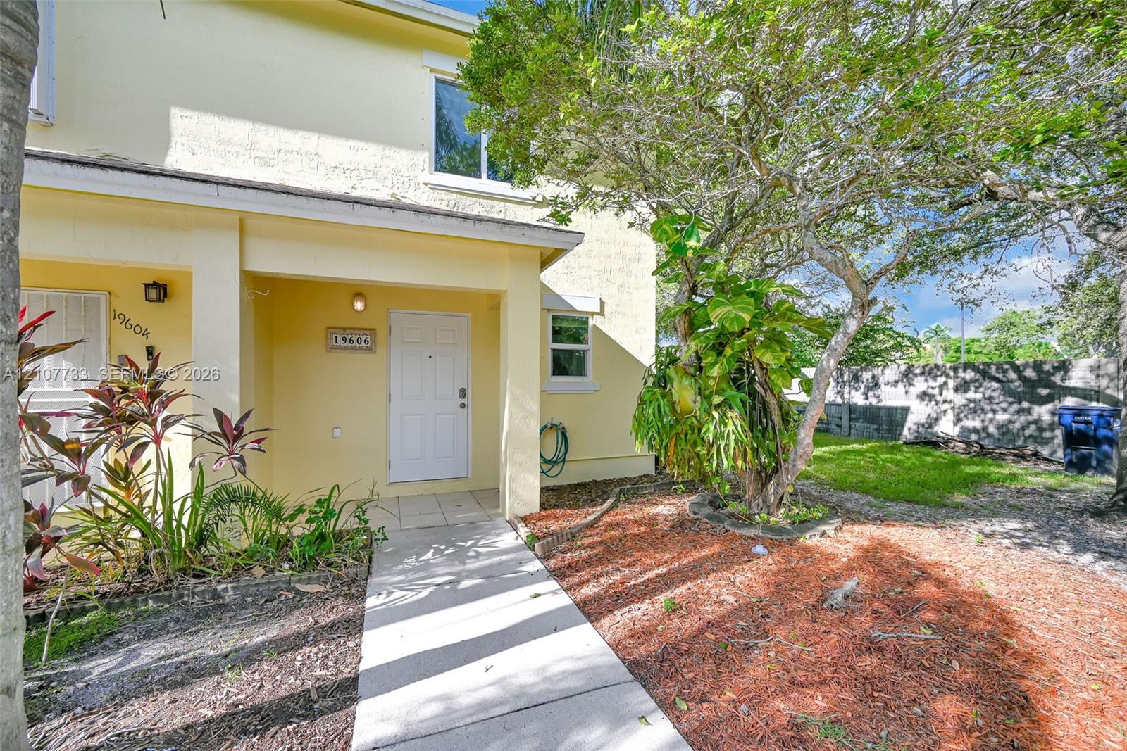 Stucco exterior featuring a covered entryway, paved walkway, mature landscaping, and an external wall light fixture