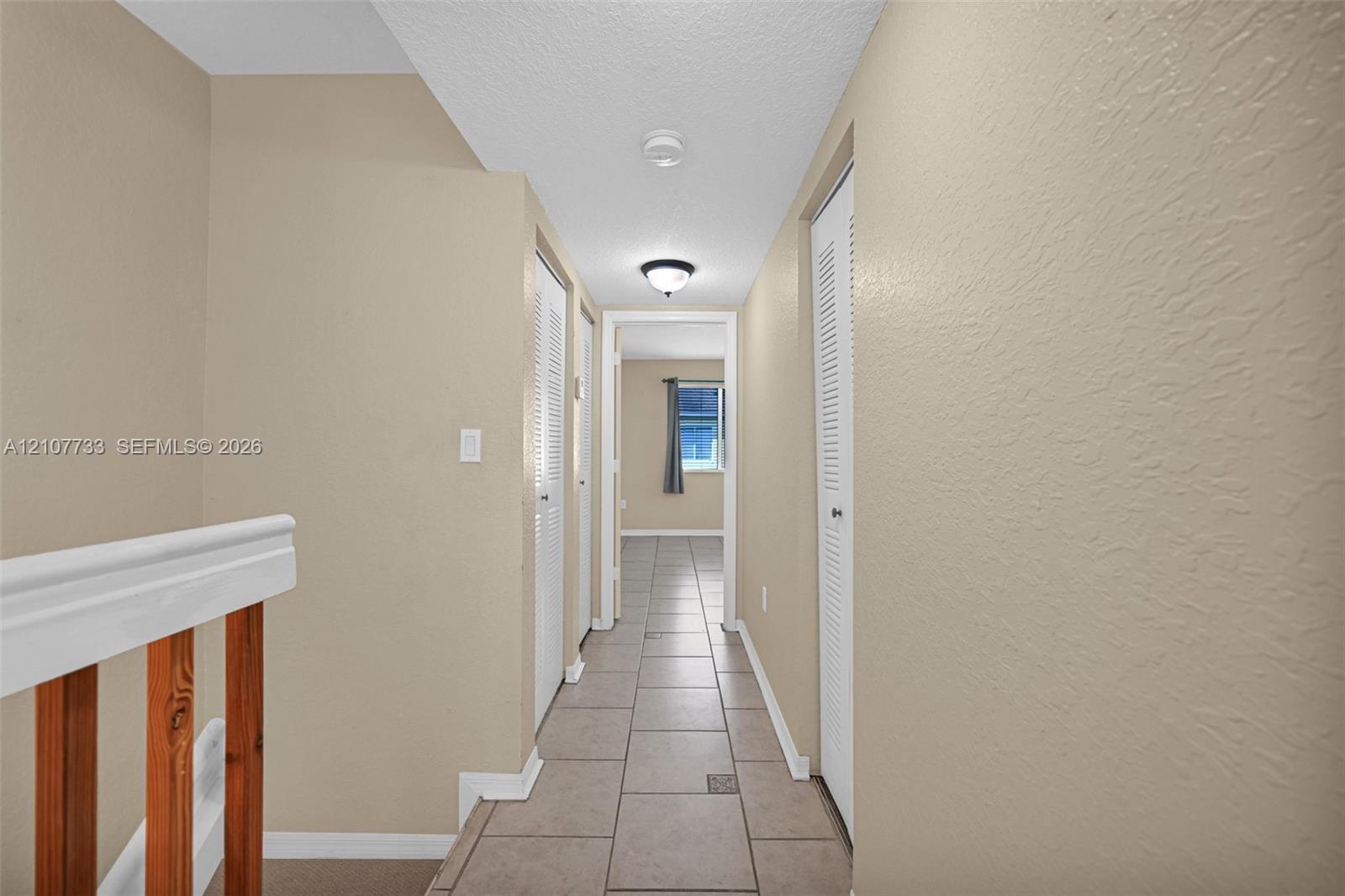 Hallway featuring large-format ceramic tile flooring, neutral wall tones, dual bi-fold closet doors, and wood-finish railing accents