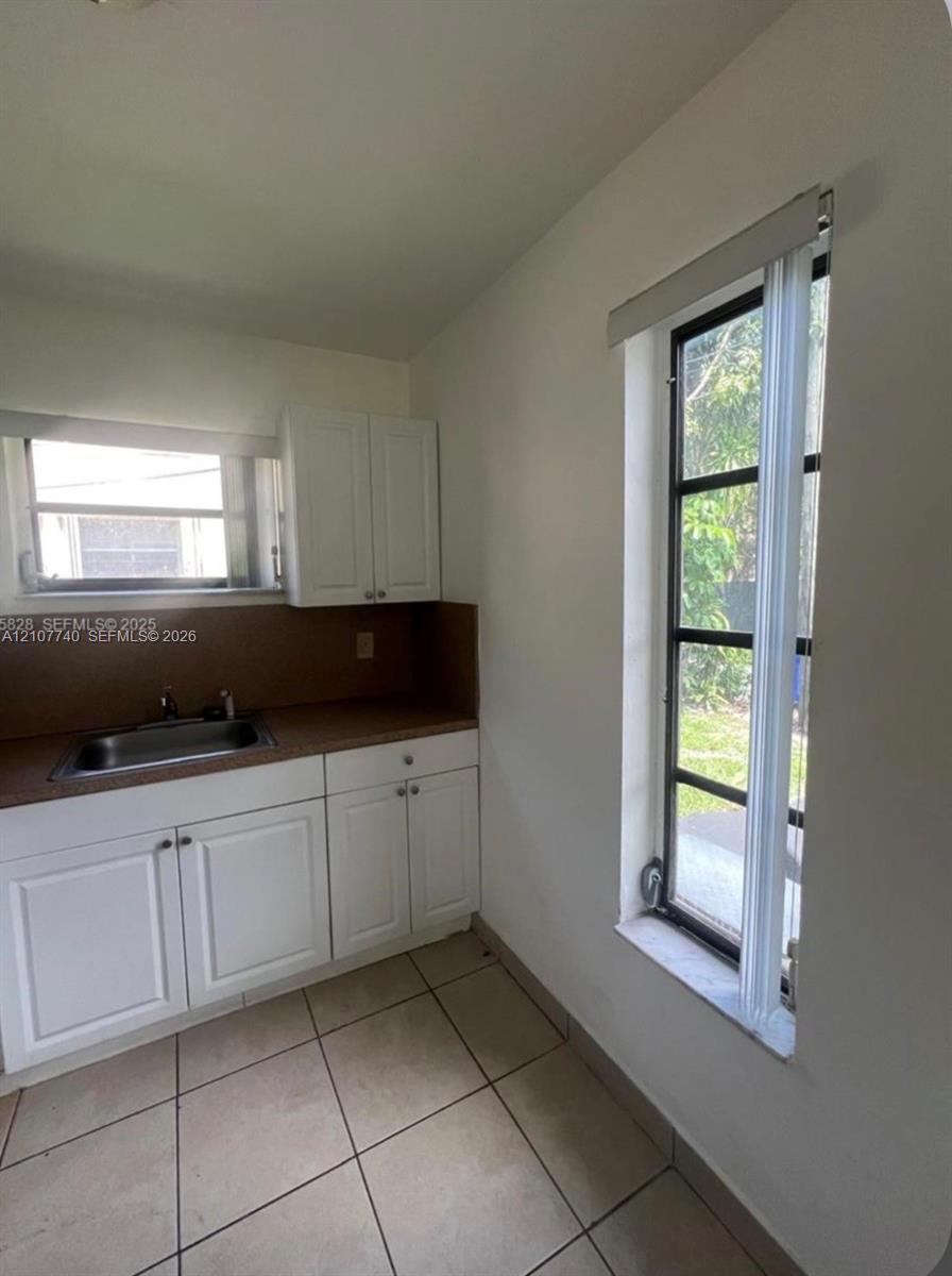 Kitchen area featuring white shaker-style cabinetry, solid-surface countertops, integrated stainless steel sink, large window with multi-pane grid, and light-toned tile flooring