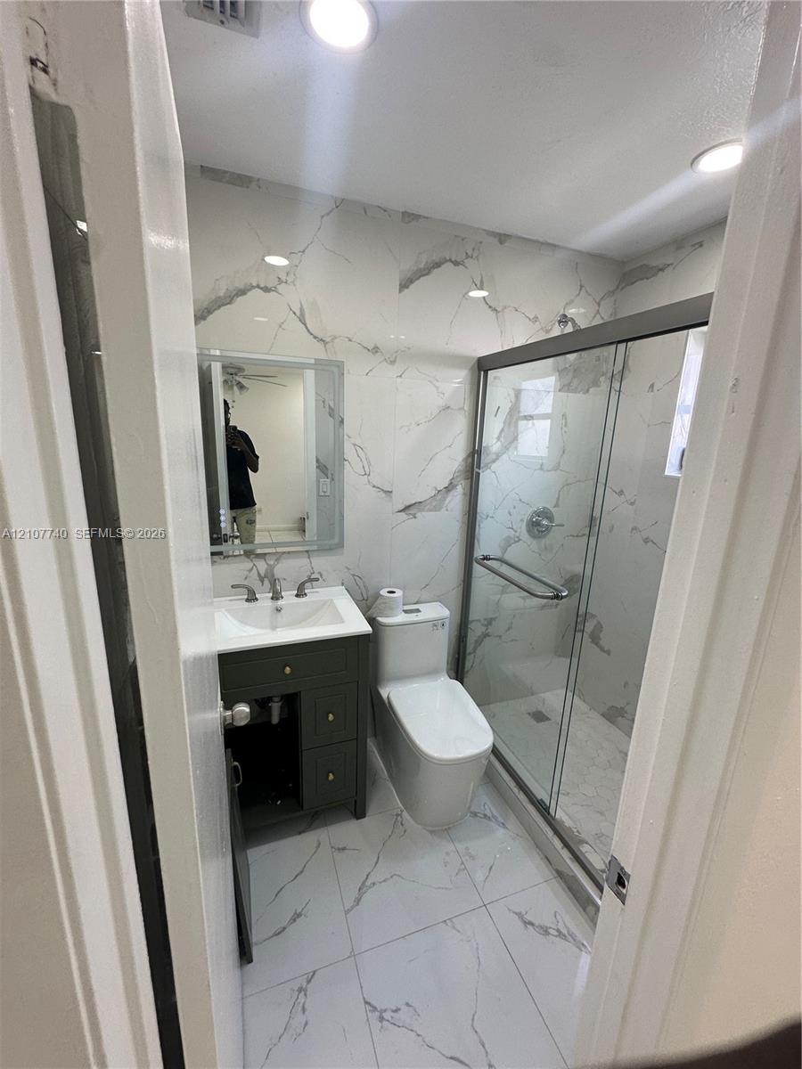 Full-height stone-finish wall tiling, glass-enclosed shower, dark-finish cabinetry with integrated sink, porcelain tile flooring, and recessed ceiling lighting