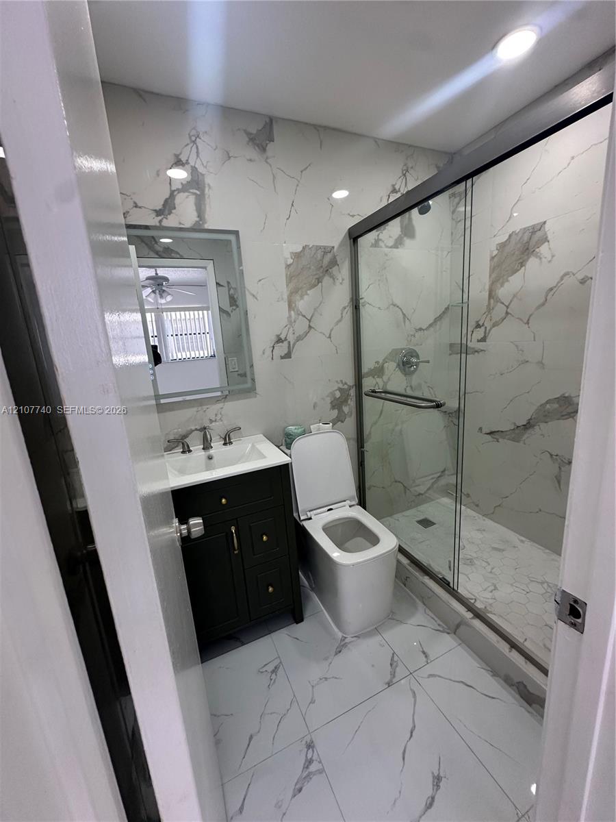 Bathroom featuring floor-to-ceiling large-format stone-patterned tile, walk-in glass-enclosed shower, dark-finish cabinetry with metallic hardware, and backlit mirror