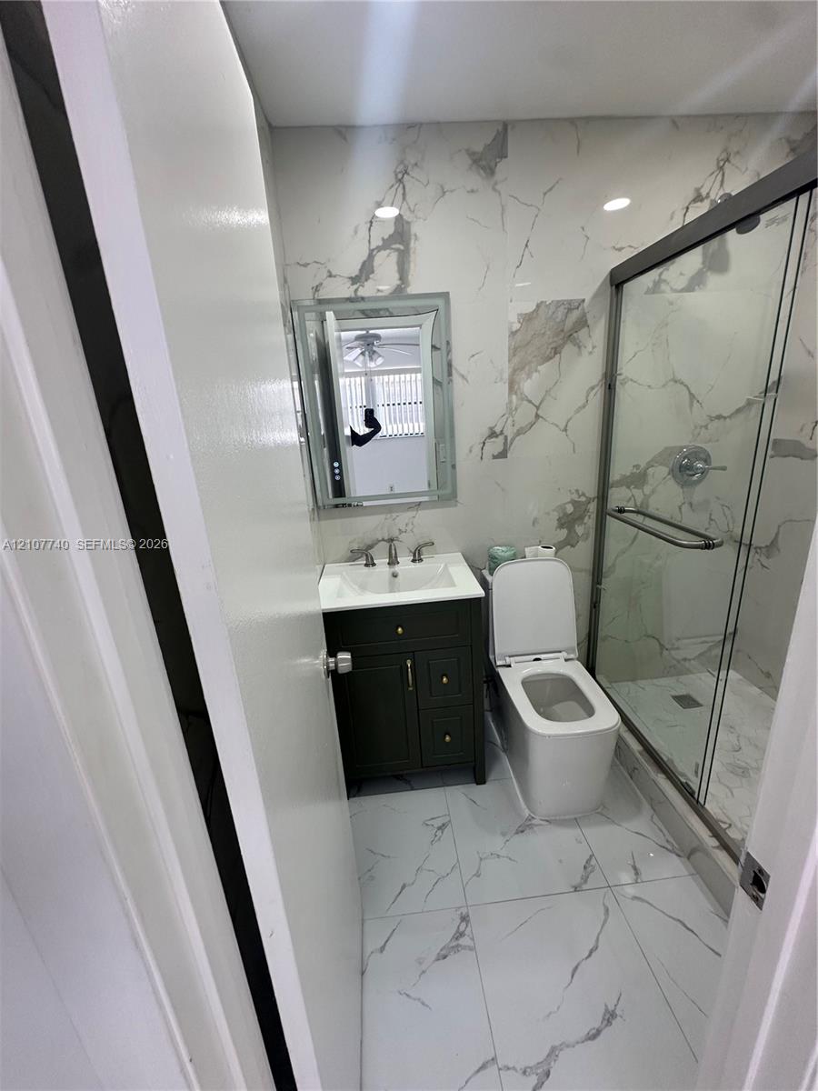 Full-height stone-patterned wall tiling, glass-enclosed shower with metallic hardware, dark-finish wood vanity with integrated sink, and large-format porcelain floor tile