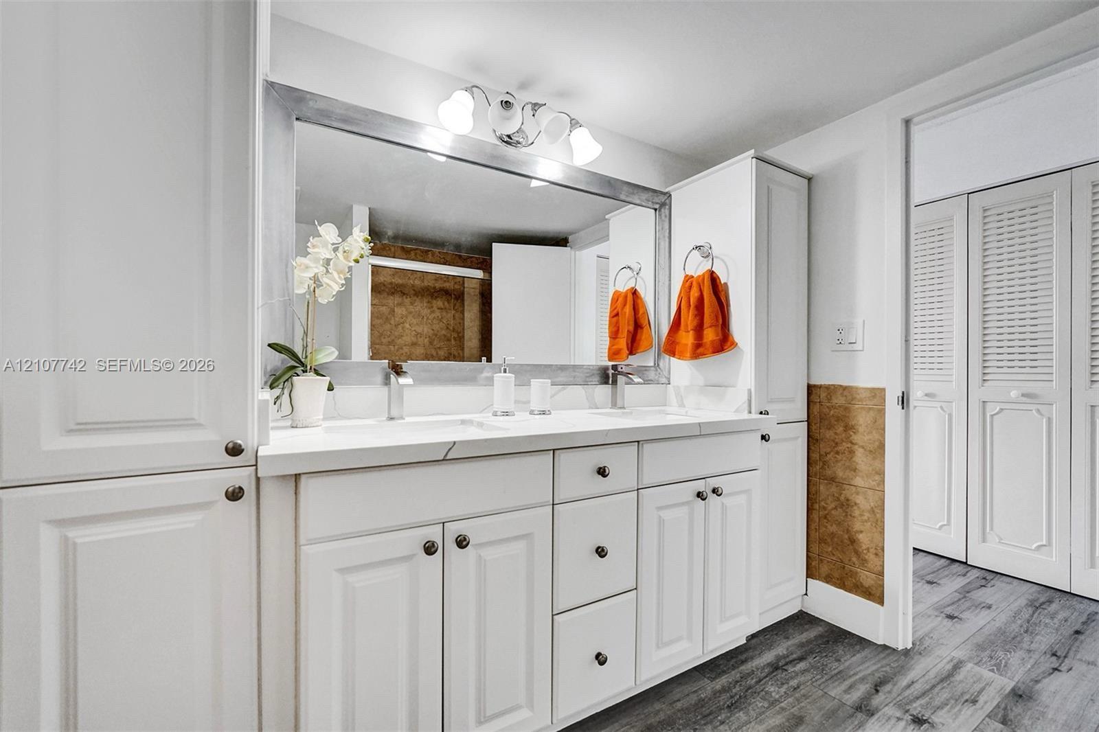Dual-sink vanity with white cabinetry, solid surface countertop, large framed wall mirror, multi-bulb overhead lighting, and white louvered closet doors