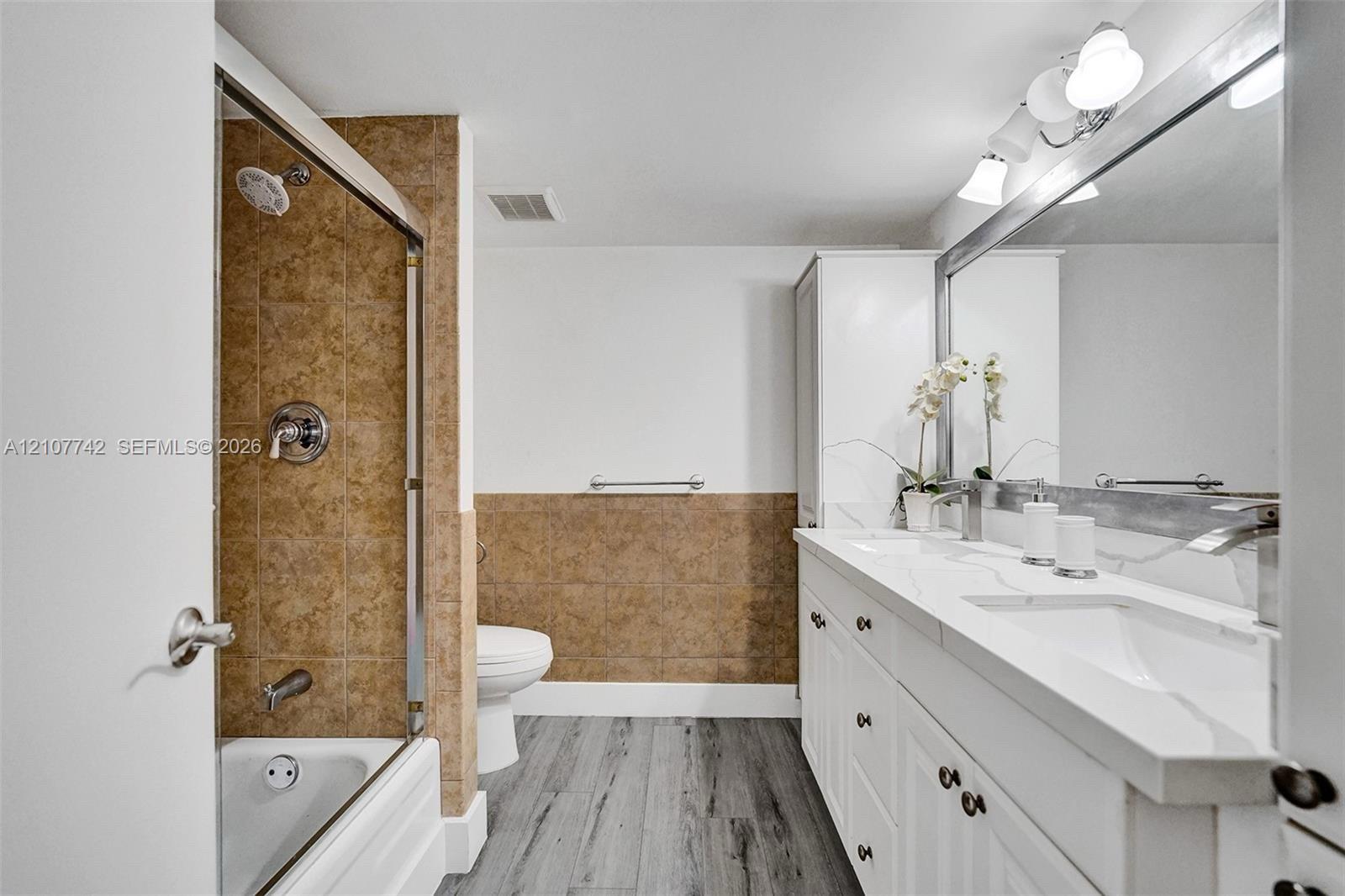 Bathroom featuring a dual-sink vanity with a solid-surface countertop, full-width mirror, shower-tub combination with tiled surround, and wood-finish flooring