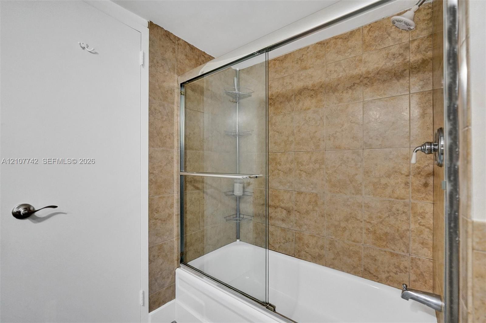 Bathtub with a glass sliding enclosure, metallic fixtures, and tiled wall surround featuring built-in corner shelving