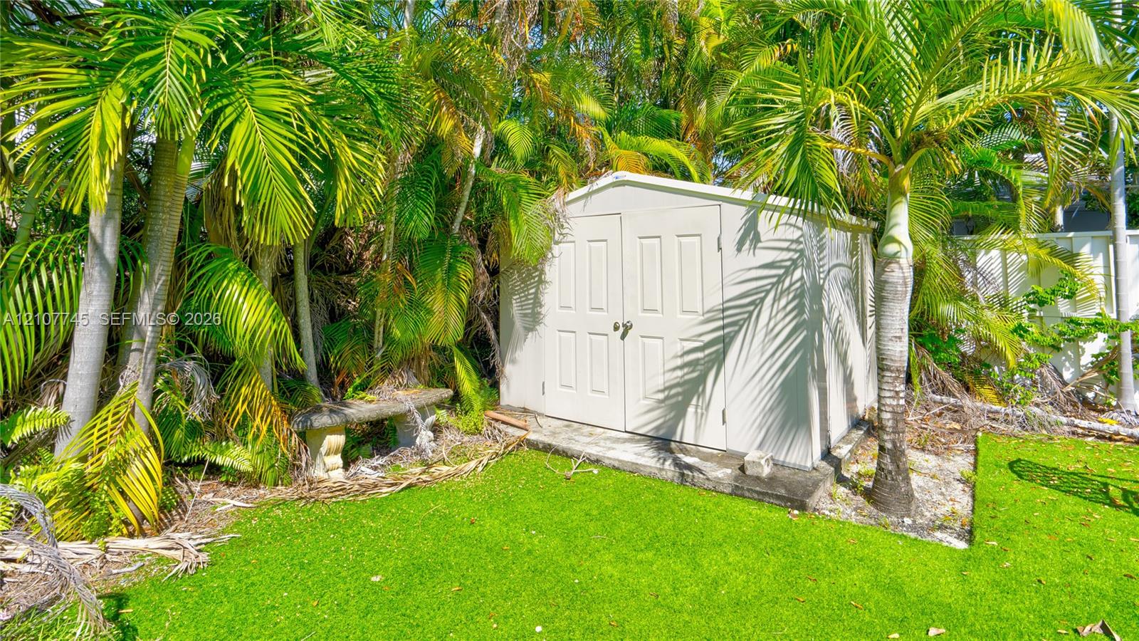 Exterior storage shed featuring double doors and a concrete foundation pad, complemented by mature palm trees and stone bench seating