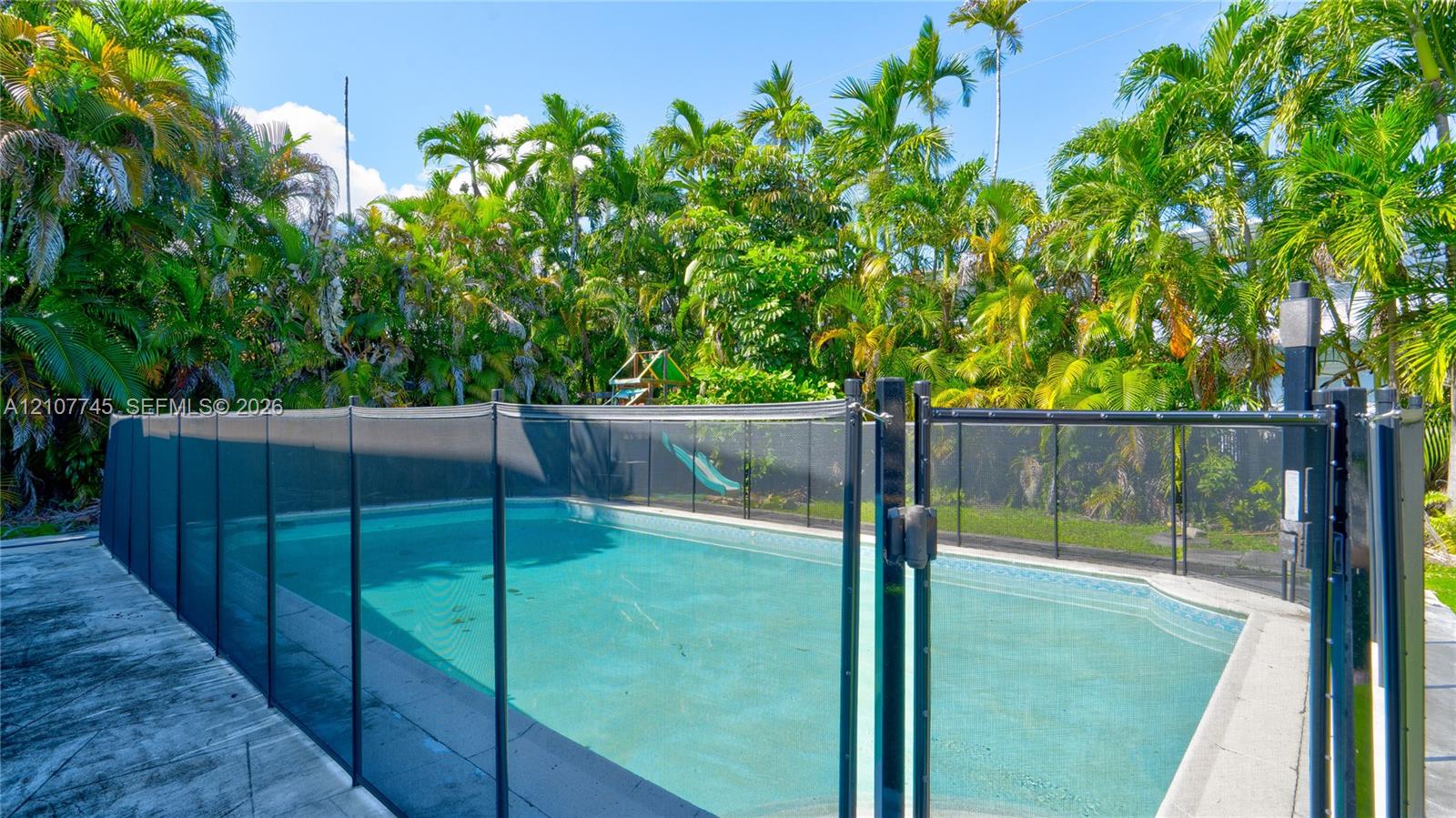 In-ground swimming pool featuring a mesh safety fence, concrete decking, and mature palm tree landscaping