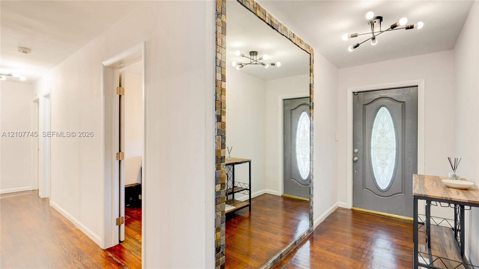 Entryway featuring wood-finish flooring, a large wall-mounted mirror with mosaic tile trim, a grey exterior door with a decorative oval glass insert, and modern multi-arm light fixtures