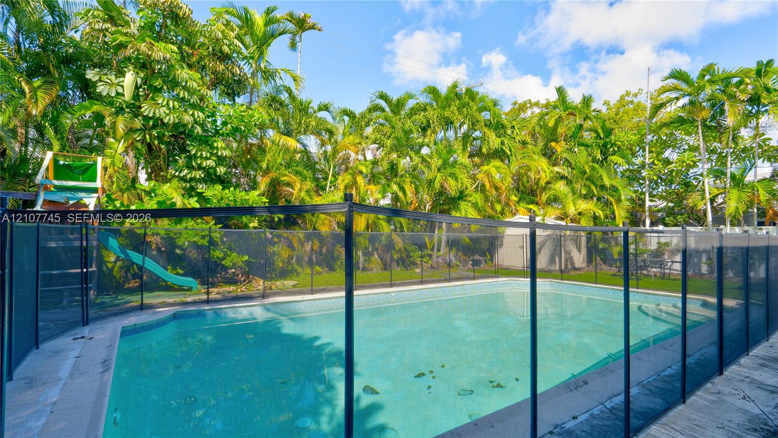 In-ground pool with integrated steps, enclosed by a black safety fence, featuring a paved deck and mature perimeter landscaping