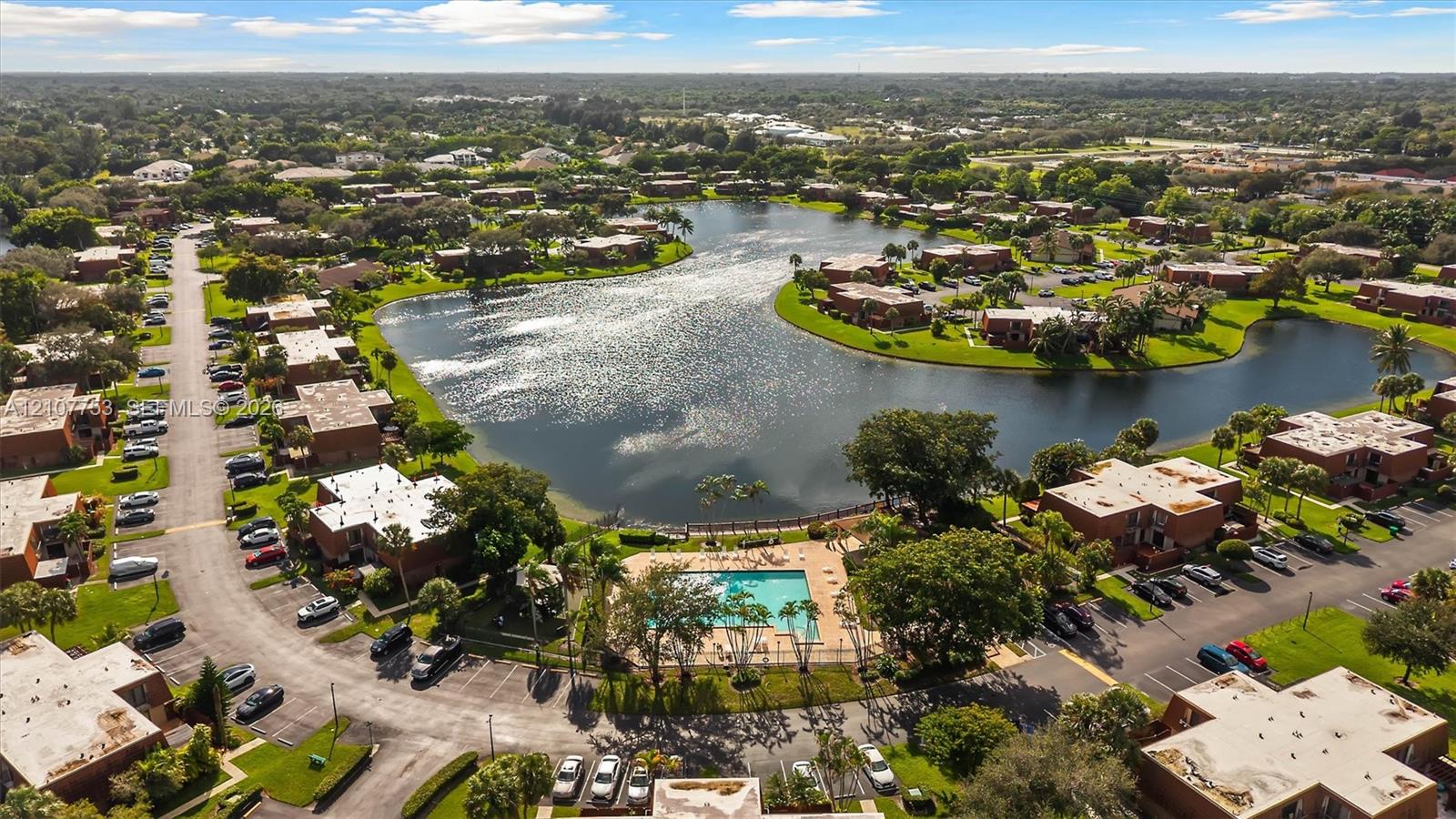 Aerial perspective of a multi-unit residential community featuring a central lake, a swimming pool with a paved deck, established trees, and asphalt thoroughfares