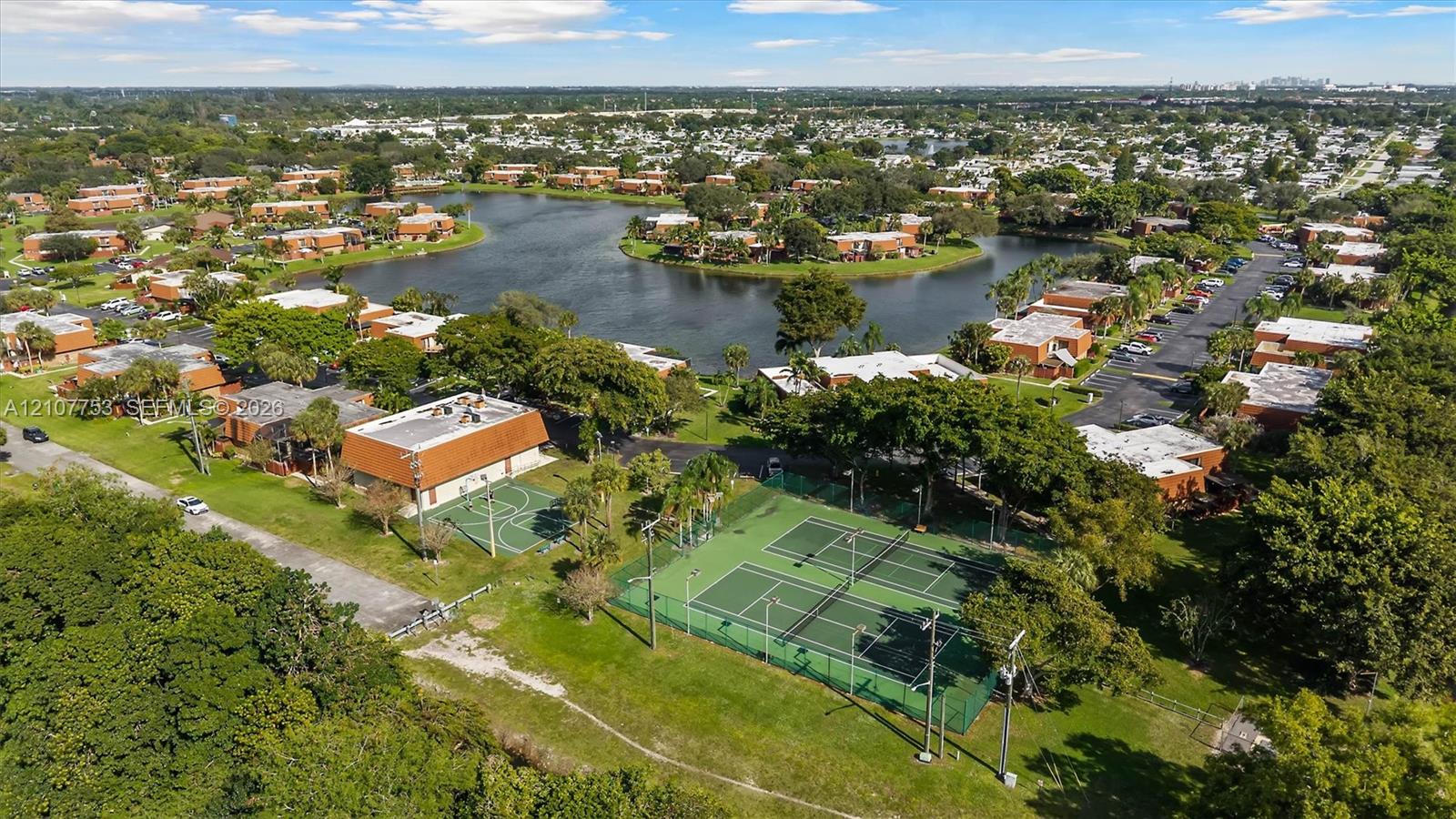 Aerial perspective of the community featuring dual tennis courts, a basketball court, surrounding water features, and mature landscaping